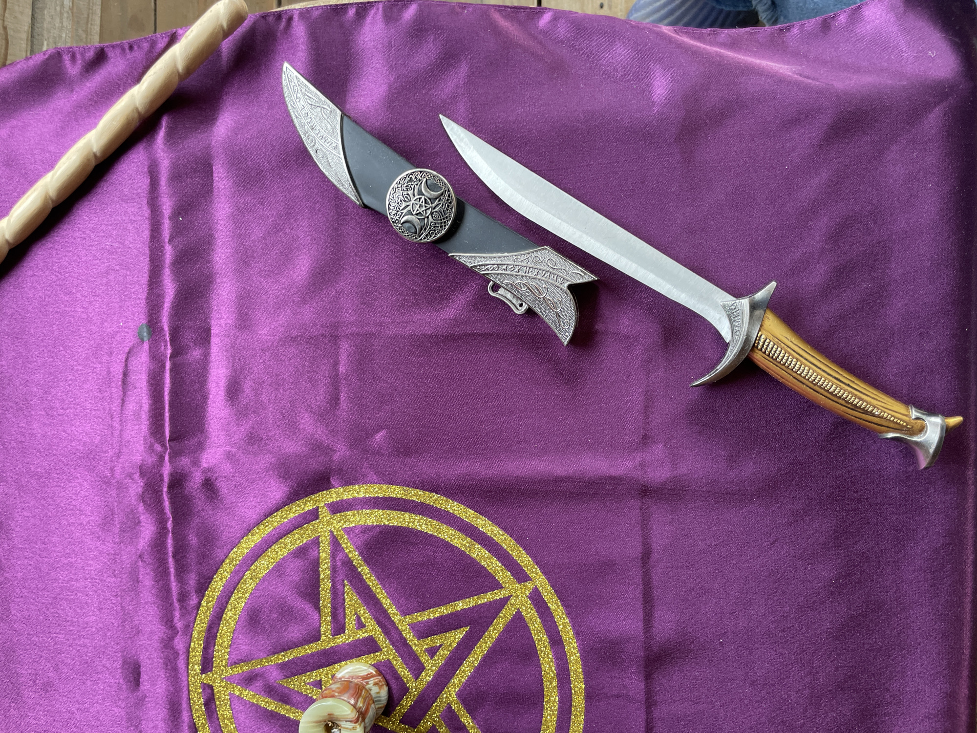 The image depicts a ritualistic scene, likely indoors. A purple cloth with a gold glitter pentagram design occupies the lower portion of the frame. A wooden staff rests on the upper left of the cloth. A dagger with a bone-colored handle and a silver bolster lies next to its black sheath, also with silver decoration, on the upper right of the cloth. There are no people visible in the image. The location of the image is unknown; Zav'yalovo, Russia cannot be determined.