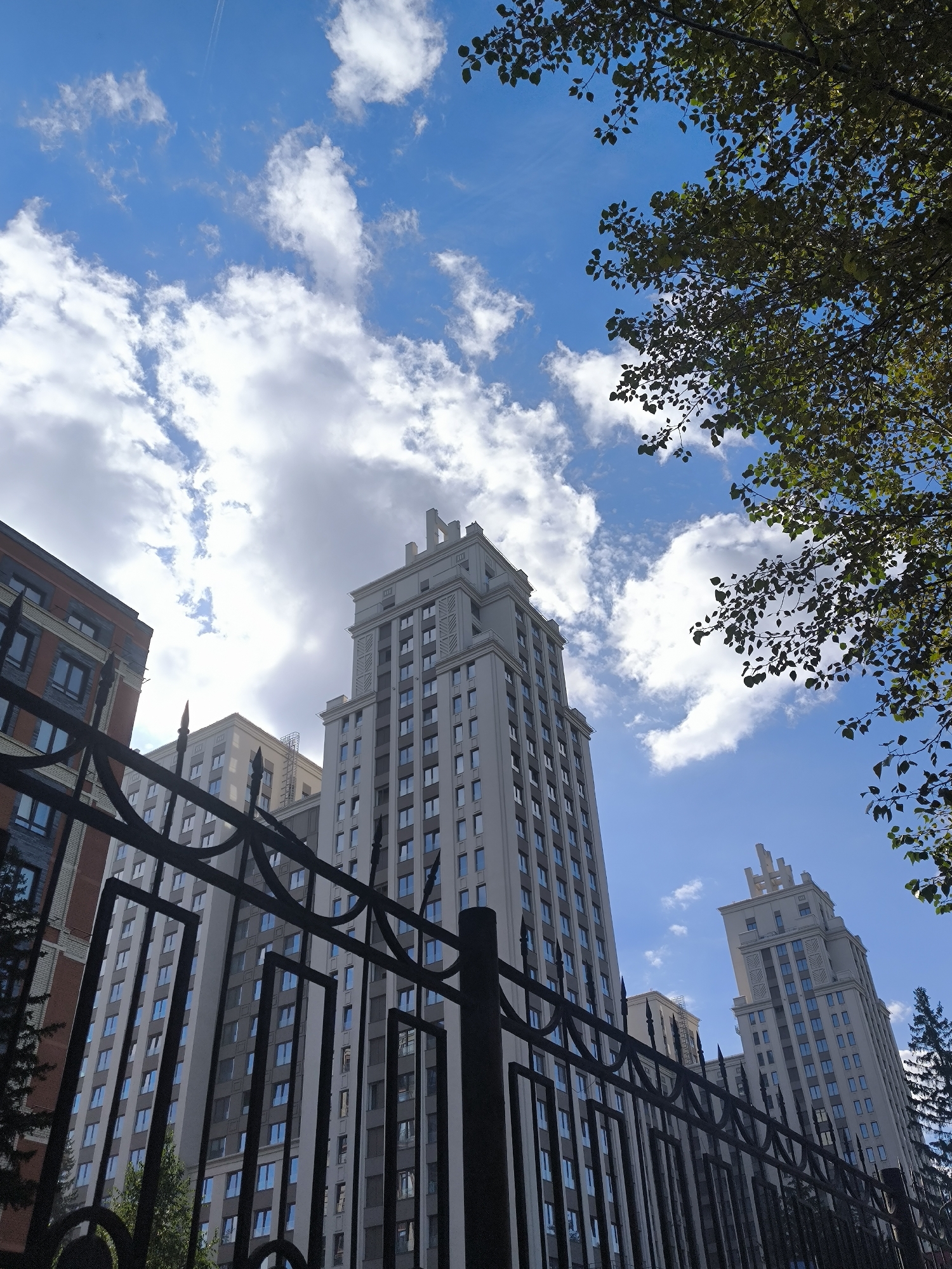 This low-angle shot captures a clear blue sky with scattered white clouds. In the foreground, a dark, ornate metal fence is visible. Behind the fence, several tall, modern apartment buildings rise skyward. The building directly behind the fence, on the left, has a reddish-brown brick facade with multiple windows. To its right, a larger, light-colored apartment building dominates the center of the frame, characterized by its numerous windows and decorative top. Further to the right, another similar light-colored building is partially visible. The branches and leaves of a mature tree overhang the scene from the top right corner. The image appears to have been taken on a sunny day. This scene is characteristic of the architectural style found in the area around the Novosibirsk State University campus in Novosibirsk, Russia, specifically near the university's main buildings and surrounding residential complexes.