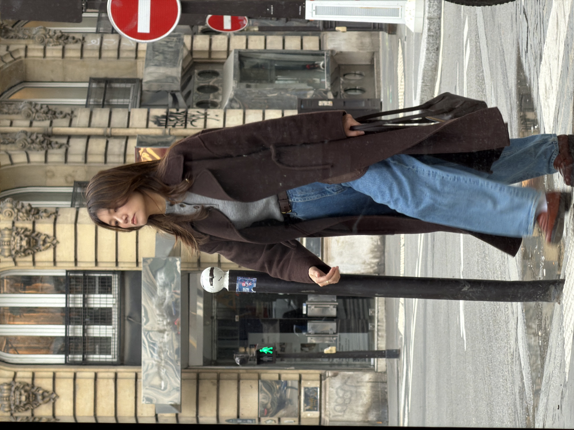 A woman wearing a brown coat, blue jeans, and brown shoes is walking across a street in Paris, France. She is carrying a brown bag. The background includes Parisian buildings, a crosswalk, street signs, and a traffic light.