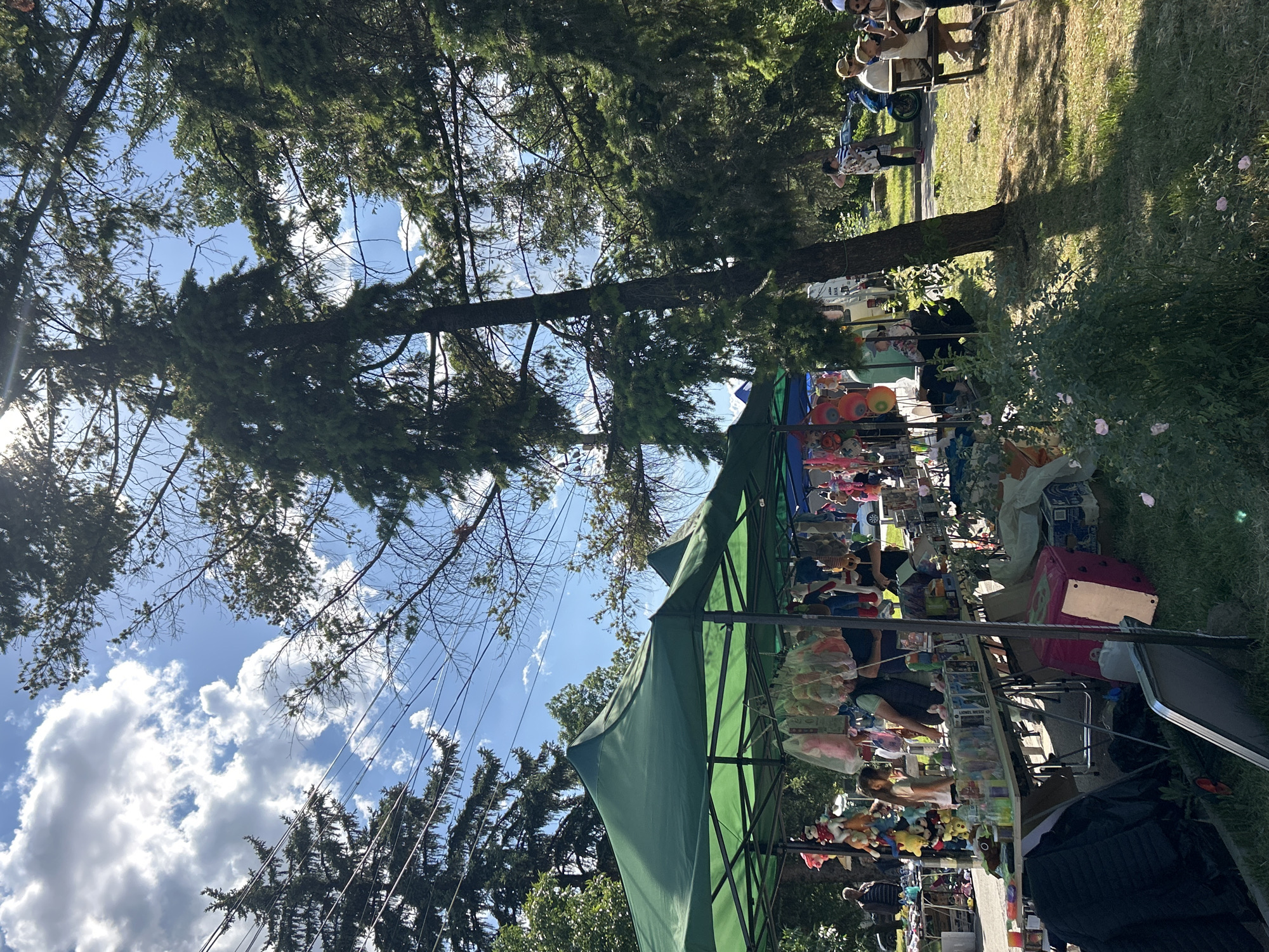The image depicts a marketplace scene in Mirovyane, Bulgaria. A green canopy tent dominates the foreground, shielding various merchandise, including stuffed animals and other items. People are visible under the tent, presumably vendors and customers. In the background, there are trees, with some visible through the tent's framework. Above, the sky is blue with some scattered clouds. In the upper right corner, additional people are visible in the distance.