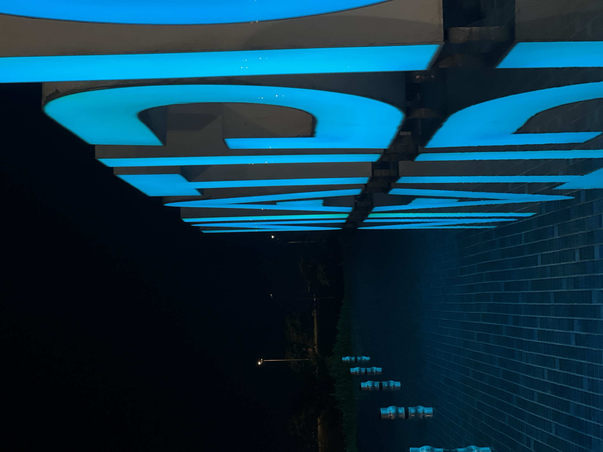 The image depicts a close-up, low-angle view of illuminated blue signage at night. The letters are block-style and appear to be mounted on a building with a brick facade. Reflected in the glossy surface of the building is a street scene with sparse vegetation and streetlights. The location is in an unknown city of unknown location.