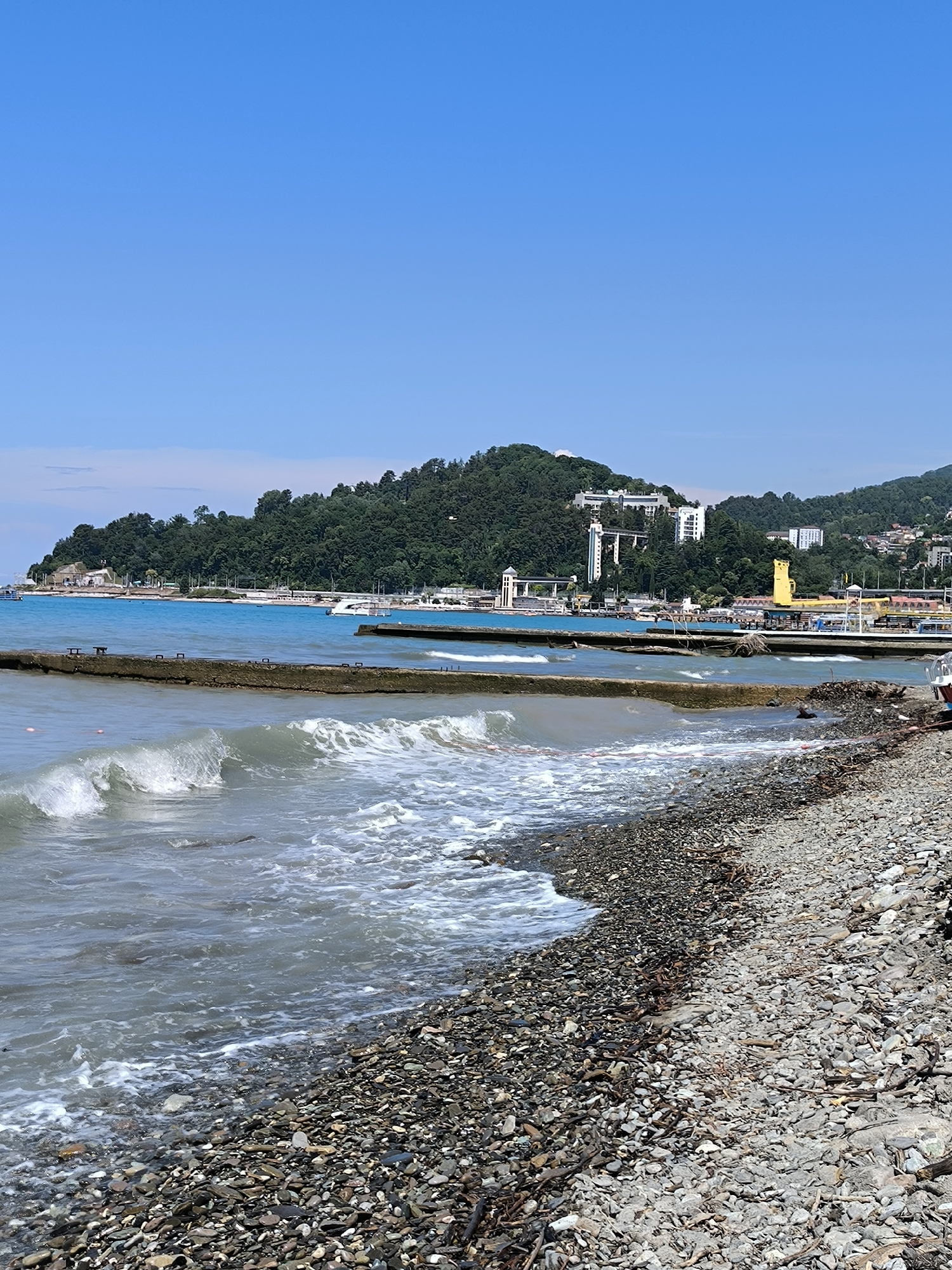 The image depicts a coastal scene in Sochi, Russia, likely a public beach area. The foreground is dominated by a pebble beach strewn with driftwood. Gentle waves are rolling onto the shore, creating white foam. Extending into the turquoise sea are concrete breakwaters, some of which appear weathered and partially submerged. In the mid-ground, the blue sea stretches towards a coastline backed by a densely forested hill. Along the coastline, there are various buildings, including white high-rise hotels, an area with a distinctive yellow structure, and what appears to be a port or promenade with boats and structures. The sky is clear and blue. There are no people visible in the image.