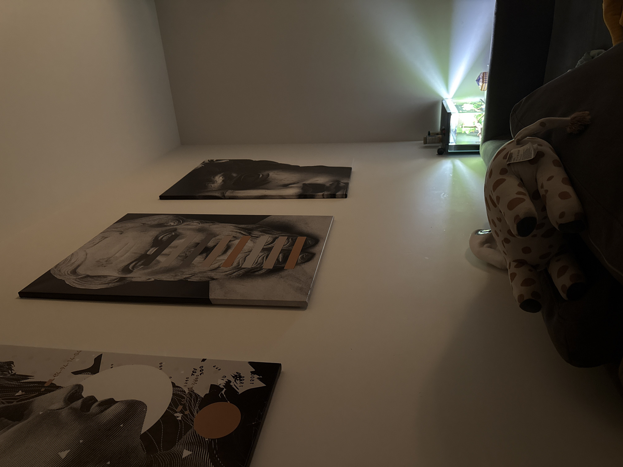 The image captures an indoor scene in Krasnoyarsk, Russia, from a low-angle perspective, looking upwards along an off-white wall. Three contemporary art canvases are mounted vertically on the wall.
The topmost canvas features a dark, abstract, wave-like form. The middle canvas displays a grayscale depiction of what appears to be a person's face, partially obscured by several rectangular bands in white, gray, and a metallic copper finish. These bands create abstract patterns, with some resembling marks like "///W". The bottom canvas shows a complex black and white graphic design that includes a human hand, abstract lines, a large white circle, and a smaller copper-colored circle. Faint text and numbers are visible on this canvas, including "53-94-14-36", "300", "004", "005", "DEC", and "QUAT".
To the right, a dark, plush sofa or couch is partially visible, with a light-colored, spotted plush toy (resembling a giraffe or deer) draped over its armrest, showing a white tag on its back. In the background, a bright, focused light source, possibly from a small aquarium or projector, casts prominent rays of light upwards and across the wall and ceiling, indicating the scene is set during evening or night. The overall atmosphere is dim, with the artificial light creating a strong visual focal point. No activity is taking place; the scene is static, showcasing the room's decor and lighting.