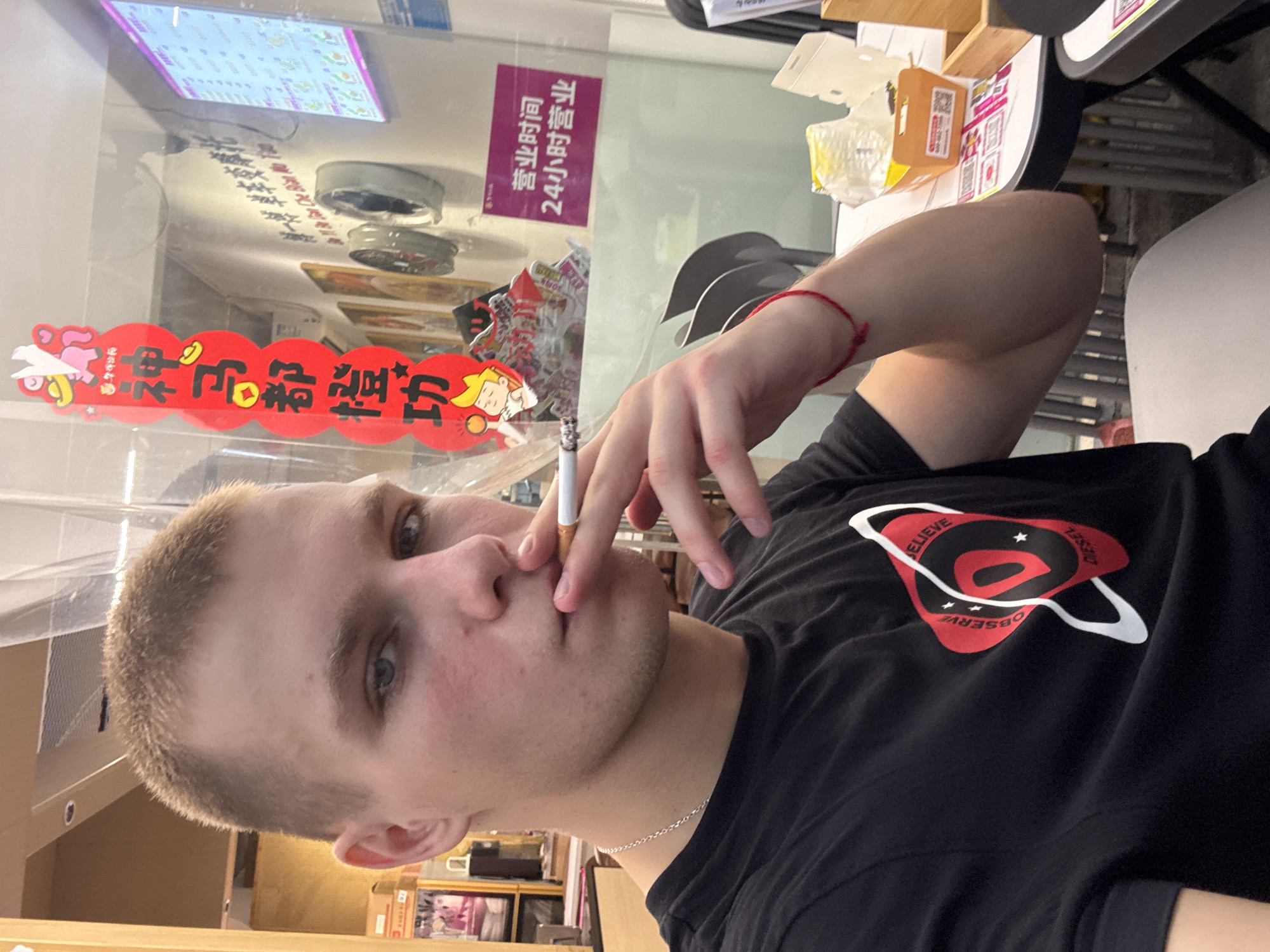 The media file shows a close-up, tilted shot of a young person with short, light-brown hair and blue eyes. They are holding a lit cigarette to their lips and appear to be smoking. The person is wearing a black t-shirt with a circular graphic on it that contains the words "BELIEVE," "OBSERVE," and "DIESEL." Around their left wrist, they are wearing a thin red string bracelet.
The background suggests an indoor setting, possibly a shop or restaurant. To the left, there is a transparent partition, possibly glass, with some red and yellow decorations featuring cartoon characters and Chinese text. One piece of text is in a purple rectangle and reads "营业时间 24小时营业," which translates to "Business hours 24 hours business." Further in the background, shelves with various items are visible. To the right, a wooden shelf unit is present, holding some stacked items, one of which is an orange box with Chinese text and a QR code.
The lighting appears to be artificial, typical of an indoor commercial space. No specific time of day or weather conditions can be determined. The overall mood is casual, with the individual focused on smoking. The location cues, such as the Chinese text and the style of decorations, point to a business in Haizhu District, China.