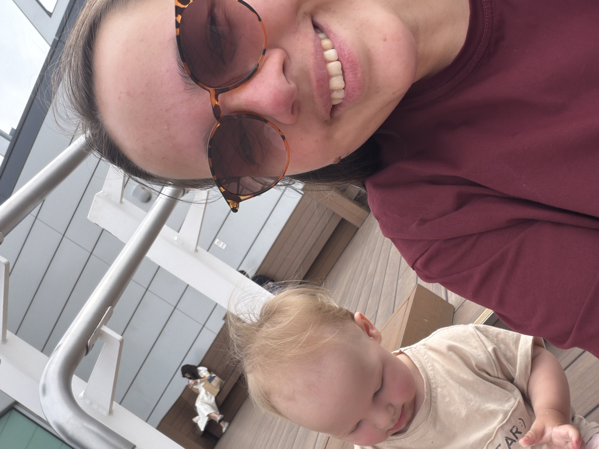 A medium shot captures a woman with sunglasses and a baby on a wooden deck, possibly outdoors given the light-colored building in the background. The woman, in the foreground on the right, wears a maroon shirt and tortoiseshell sunglasses, smiling at the camera. Her hair is dark brown. The baby, to the left, wears a light-colored t-shirt and is looking down. The background shows a woman in a white outfit sitting on a bench and a man walking in the distance. The scene appears to be in the city of Ota, Japan, likely at a public space or a commercial area with an outdoor seating section.