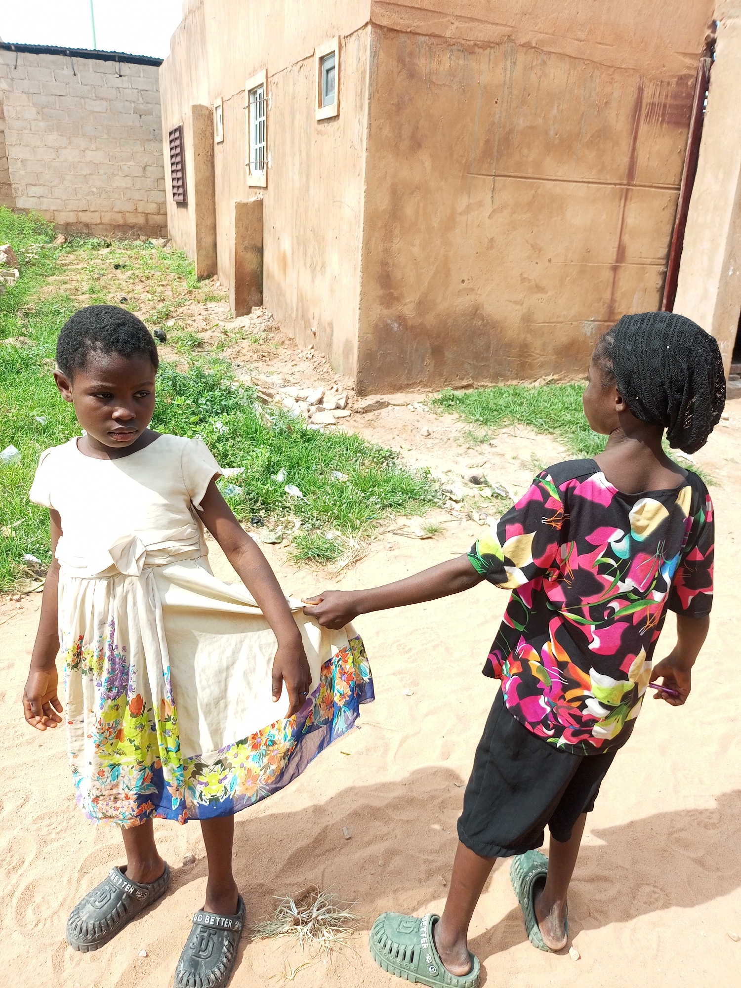 Two young girls stand on a sandy path in front of a tan-colored, mud-brick building. The girl on the left, wearing a cream-colored dress with a floral hem and dark gray Croc-style shoes, looks towards the camera with a serious expression. The girl on the right, wearing a black headscarf, a colorful floral print t-shirt, black shorts, and green Croc-style shoes, is pulling on the hem of the other girl's dress. The background shows a section of the mud-brick building with a barred window and a smaller window. To the left of the building, a lighter-colored brick wall is visible, with patchy green grass and some debris on the ground. The overall scene appears to be in a residential area of Gombe, Nigeria.