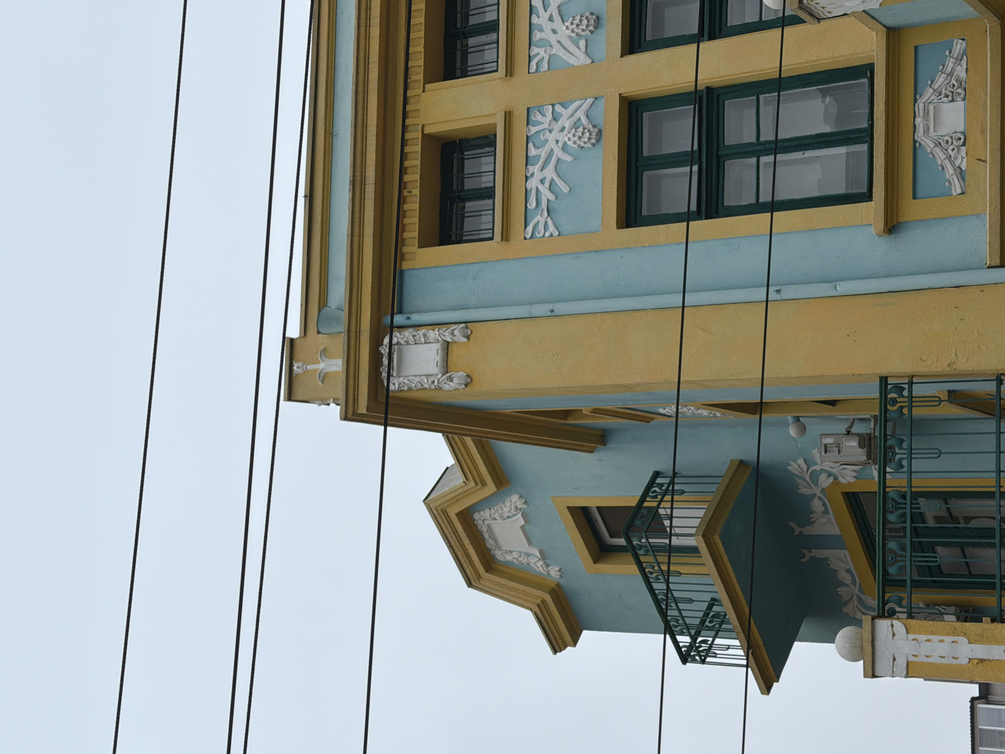 The image shows a building facade, likely located in Varna, Bulgaria. The building features a two-story structure with a combination of yellow and blue painted sections. The architectural details include framed windows with green trim, decorative white plaster elements resembling foliage, and ornate balconies with metal railings. Power lines are visible against a pale sky.