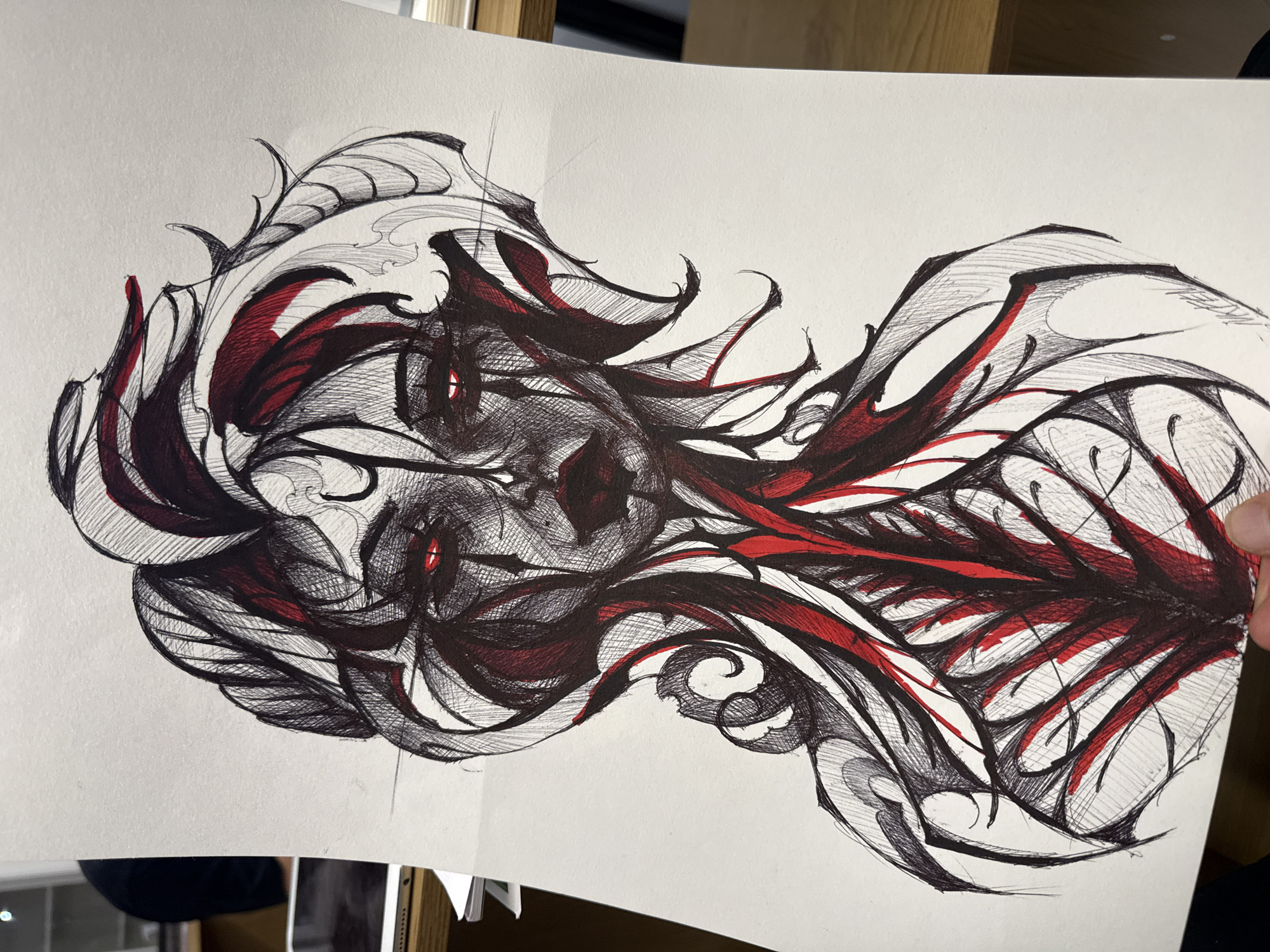 The image presents a pen and ink drawing featuring an abstract figure rendered primarily in black with red accents. The central form appears to be a stylized face with sharp angles and intense, piercing red eyes. The surrounding lines and shapes create a sense of motion and depth, evoking a surreal or fantastical atmosphere. The drawing is on white paper and appears to be held or displayed in an indoor setting, with a wooden shelf or structure visible in the background. The environment does not provide enough detail to identify a location in Sofia, Bulgaria.
