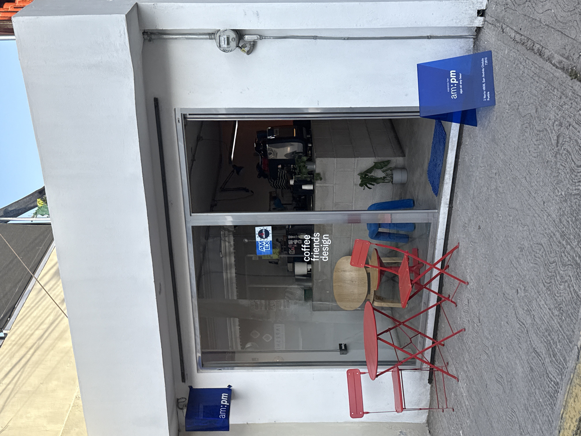 The image captures the exterior of a café named "am:pm" located in San Andrés Cholula, Puebla, Mexico. The café's façade is white, with a large glass sliding door that allows a view of the interior. A small, round wooden table and two red bistro-style chairs are positioned just inside the entrance. A blue A-frame sign placed on the sidewalk outside the entrance displays the café's name and indicates that it is an "espresso bar" open "sips at any hour," with its address at 2 Norte #808, San Andrés Cholula, 72810, Puebla. A smaller blue sign with the café’s name is attached to the building wall next to the door frame. The interior is partially visible, revealing a minimalist design with plants and what appears to be a counter area. The pavement outside is textured, suggesting concrete. A utility meter is visible on the white wall above the door. The café's name and the phrases "coffee friends design" and "AM ABIERTO EX" are displayed on the glass door.