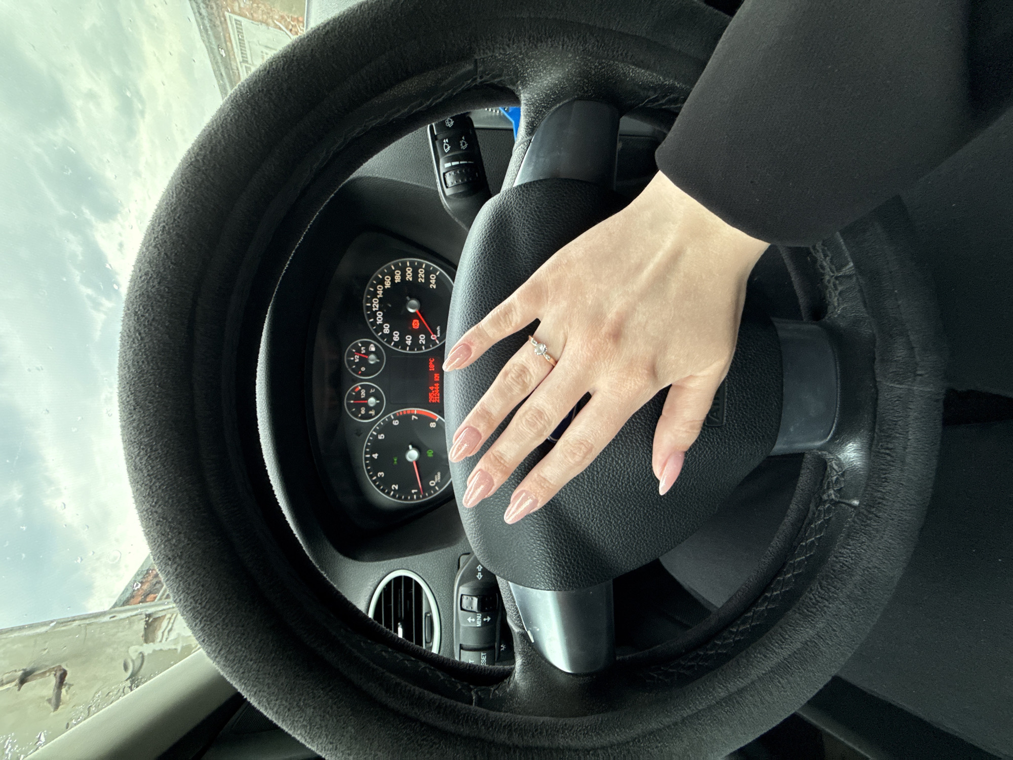 The image depicts an interior view of a car, specifically focusing on the steering wheel and the driver's hand resting upon it. The steering wheel is covered in a plush, likely aftermarket, material, and the dashboard gauges are visible behind the central portion of the wheel. A hand with manicured fingernails is prominently displayed, adorned with a ring. The person is wearing a dark-colored blazer or coat. Looking through the windshield, the outside environment includes what appears to be a rainy or overcast day, with a building visible through the upper-left part of the glass. Based on the OCR, the outside temperature is approximately 18°C. The OCR also contains the string `213444 RIV`. The location is Chaikovsky, Russia.