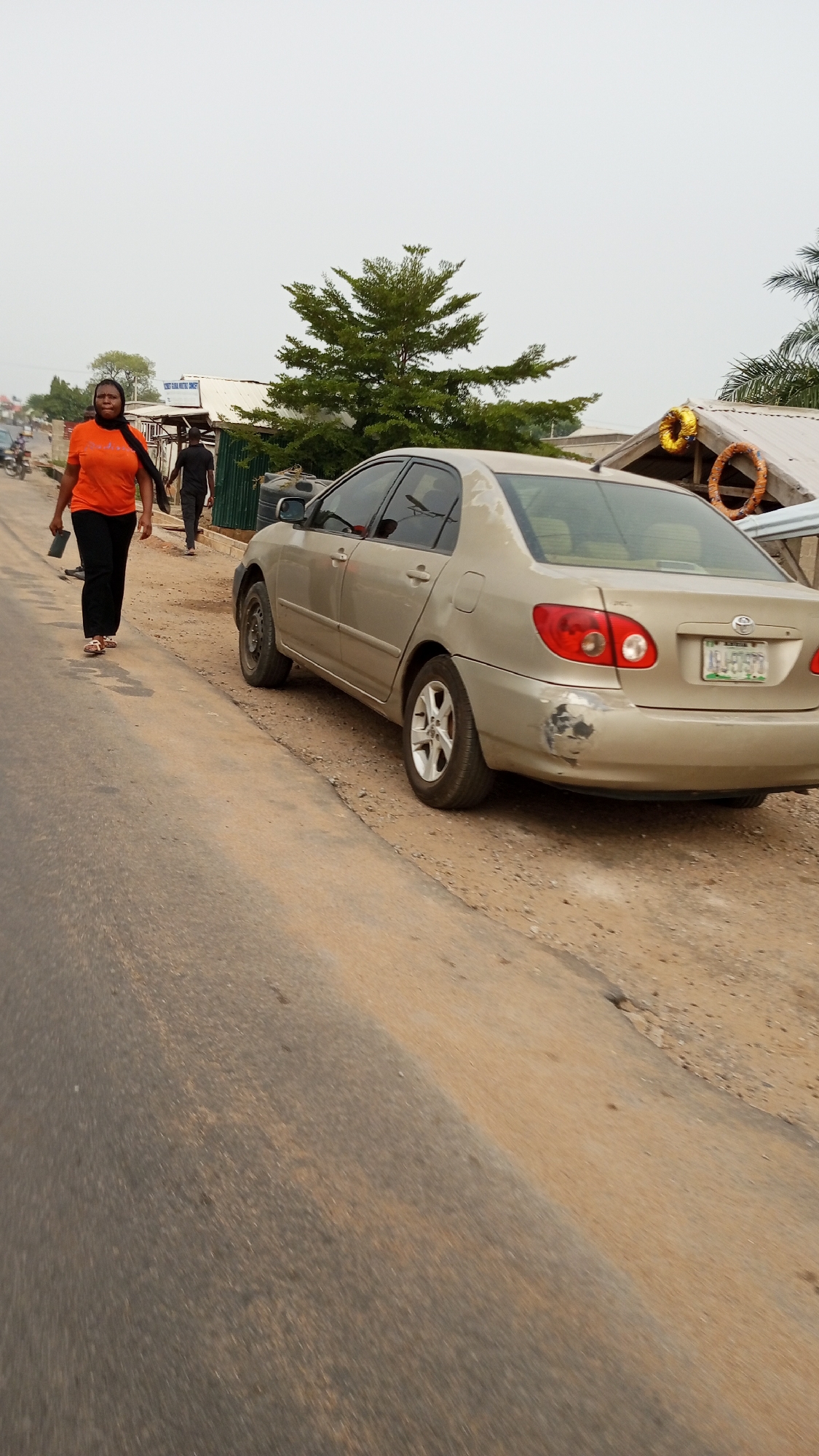 The image shows a street scene in Zango-ndaji, Nigeria. An older model gold Toyota Corolla is parked on the right side of the road. The car has damage to the rear bumper. A woman wearing an orange shirt and black pants is walking on the left side of the road. Behind her, another person is walking in the same direction. In the background, there are buildings with corrugated iron roofs and a few trees. The weather appears overcast.