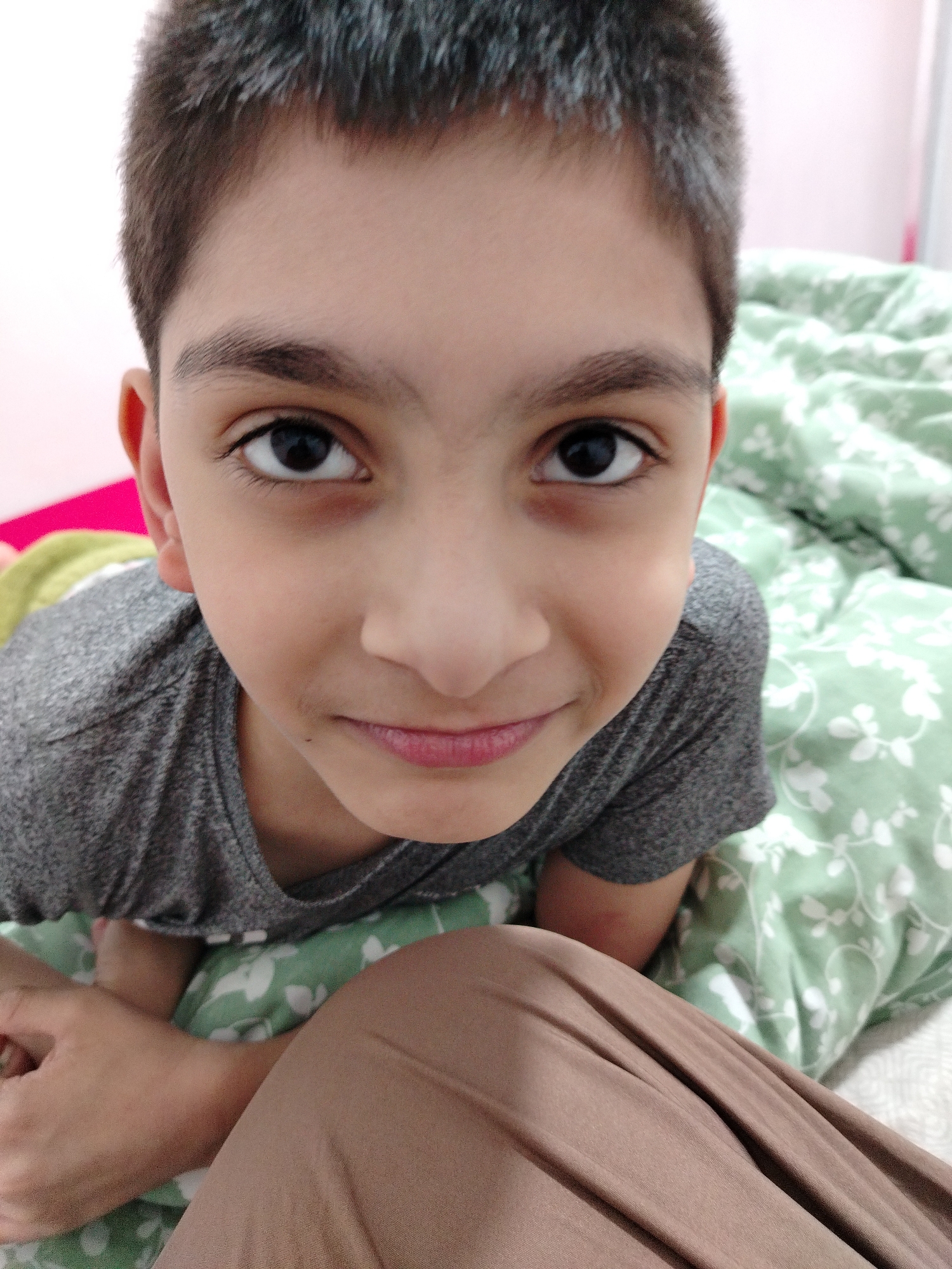 This is a close-up, eye-level, medium shot of a young boy lying down, looking directly at the camera. He has short, dark hair and prominent eyebrows. His eyes are dark and wide open, and he has a slight smile. He is wearing a grey t-shirt. The boy is lying on a green comforter with a white floral pattern. A portion of his legs, covered by beige pants, are visible in the foreground, as is his left arm and hand. The background appears to be a light-colored wall. The image is likely taken indoors, possibly in a bedroom. There is no discernible urban context to place it in Al Mas'oudi, United Arab Emirates.