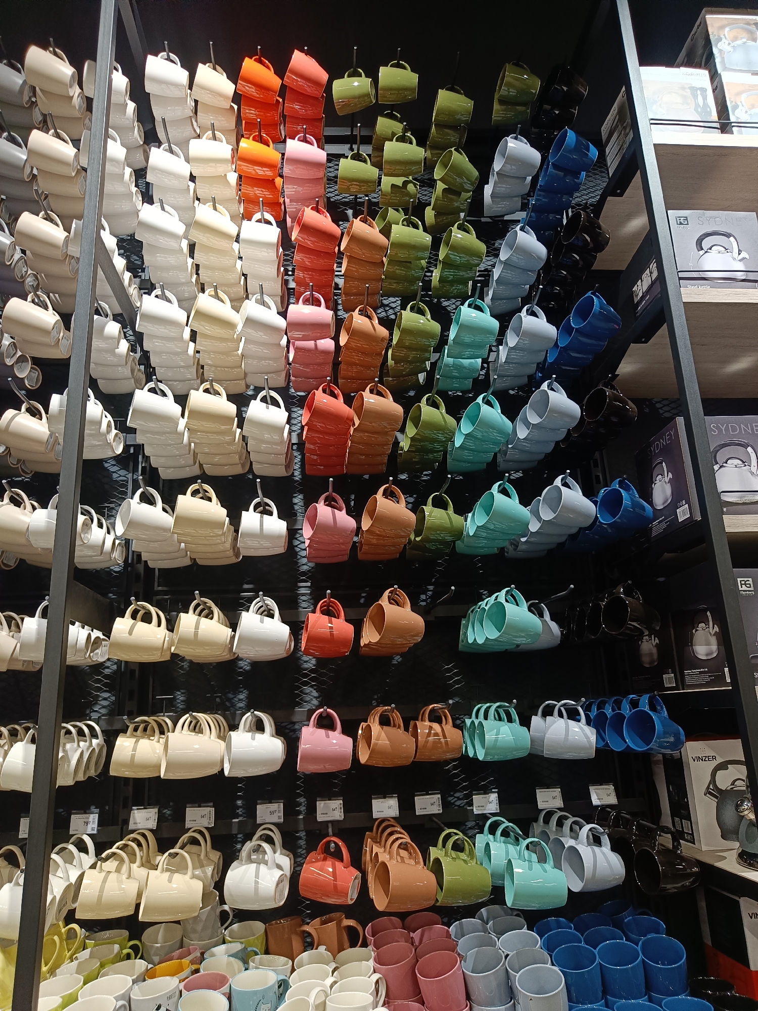 The image displays a retail store shelving unit filled with stacks of ceramic mugs. The mugs are arranged in rows and columns, with various colors including white, cream, beige, pink, orange, brown, green, teal, light blue, and dark blue. The shelves are made of dark metal, and the mugs are primarily displayed in sets of four, with handles facing outwards, suspended from metal hooks. In the background, there are boxes and packaging for kitchen appliances, including kettles. The scene is brightly lit, suggesting an indoor retail environment. No people are visible in the image.