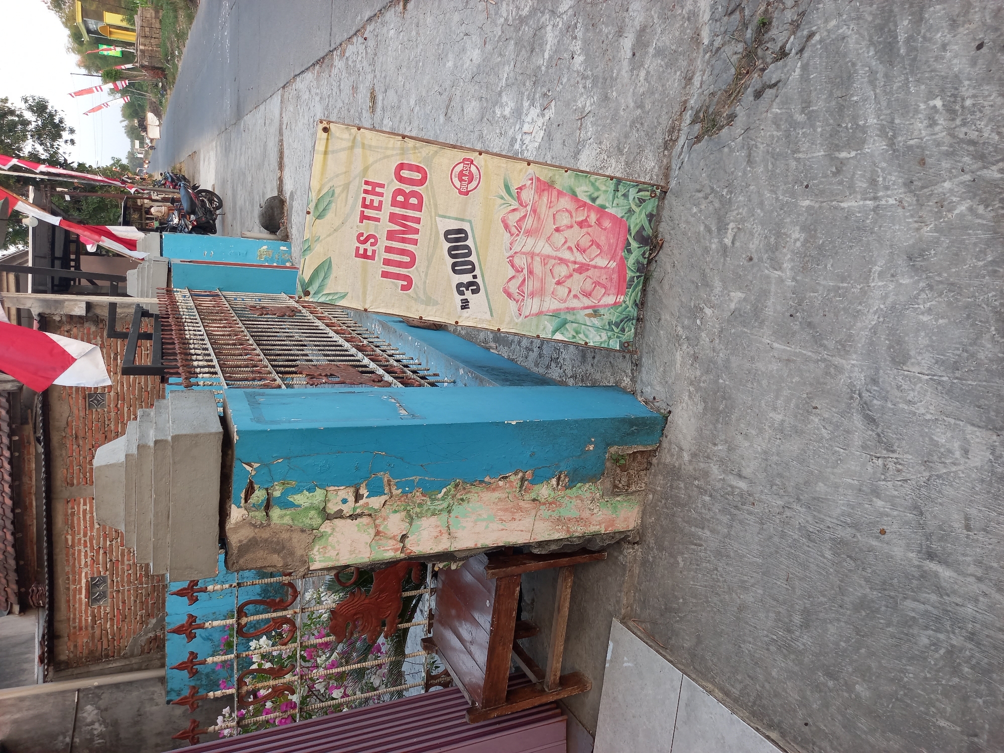 The image depicts a street scene in Kebon Agung, Indonesia, at ground level. A blue painted pillar, heavily weathered and peeling, stands prominently in the foreground, supporting a rusted metal fence. Adjacent to the pillar is a simple wooden chair. Further down the sidewalk, a yellow banner advertising "ES TEH JUMBO" for Rp 3,000 is propped up. The banner features an illustration of two glasses filled with iced tea. Behind the fence, a brick building with a tiled roof is visible, adorned with Indonesian flags. A road runs alongside the sidewalk, with several motorcycles parked and a few people in the distance. The overall environment suggests a typical roadside scene in a rural or semi-urban Indonesian setting.