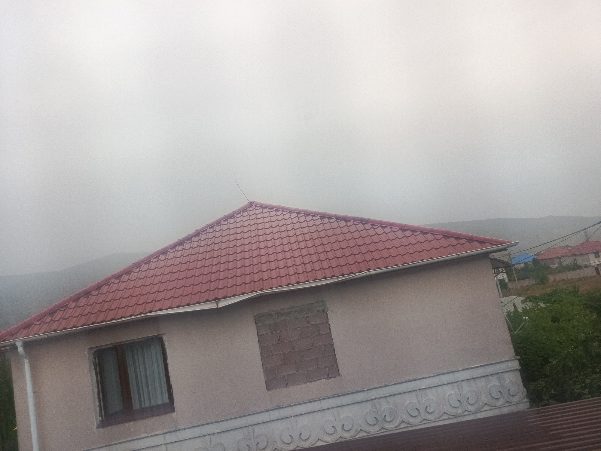 This image depicts a side view of a two-story house with a red tiled roof and light-colored walls. A single window with white curtains is visible on the first floor. A section of the wall has been bricked up. The house is situated in a rural or suburban area with rolling hills and vegetation in the background, shrouded in fog. In the distance, other houses with red roofs can be seen. The weather appears overcast and foggy. The image does not contain any people or indicate specific actions or interactions.