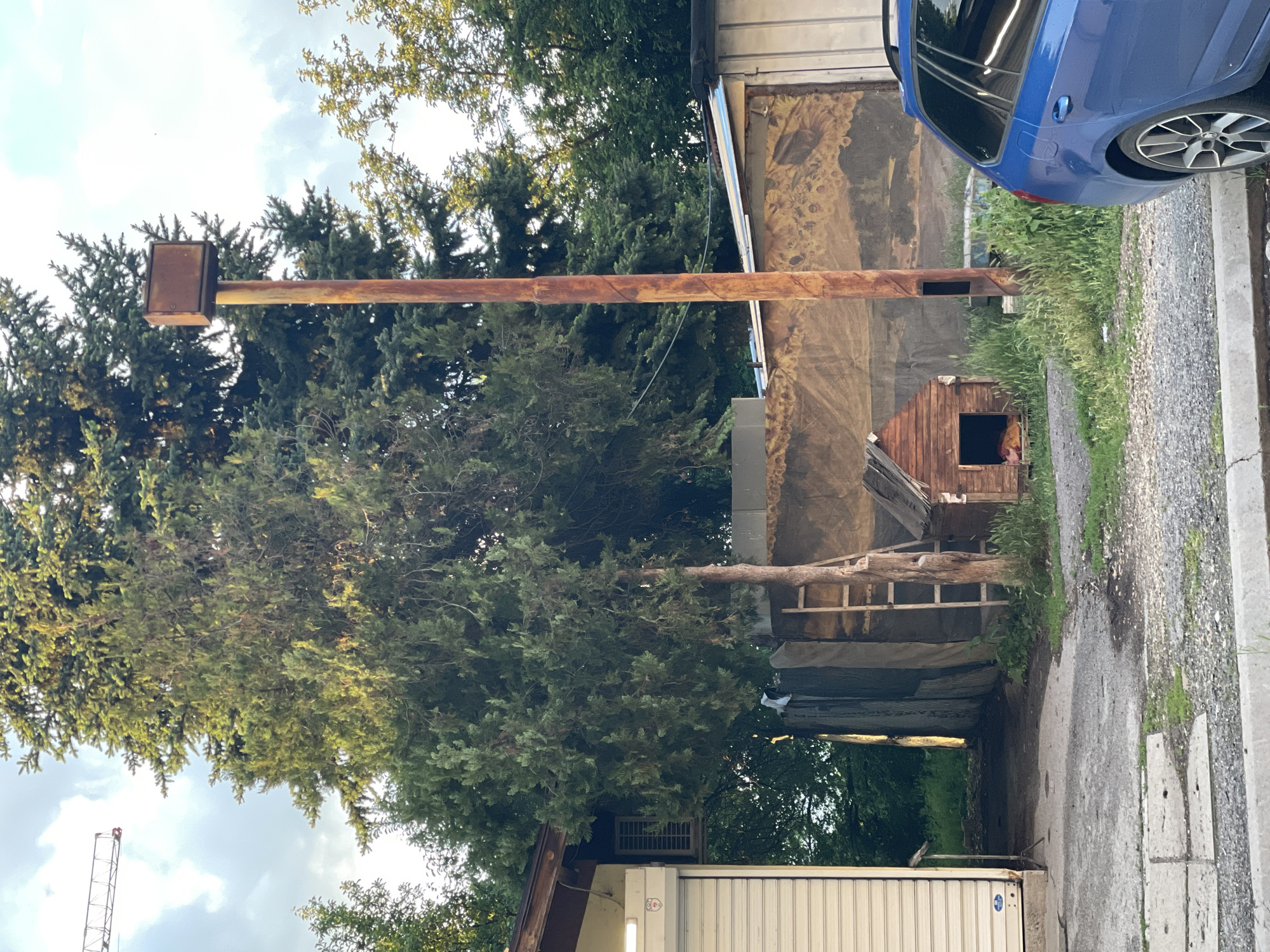 The image depicts an outdoor scene. A rusty metal pole extends horizontally across the frame, with a metal box at its left end. A tree grows in front of the pole. A blue car is parked on the right side of the frame, partially visible. Below the pole and the car, a small wooden dog house sits on a gravel driveway. A building with an elaborate mural on the wall is present in the background. Additional building structures and trees are visible in the background. A crane is visible in the lower left corner. Based on the architecture and overall appearance, the location appears to be in a rural or semi-industrial area on the outskirts of Sofia, Bulgaria, given the Eastern European architectural style and the presence of graffiti art, as well as industrial style storage in the lower right.