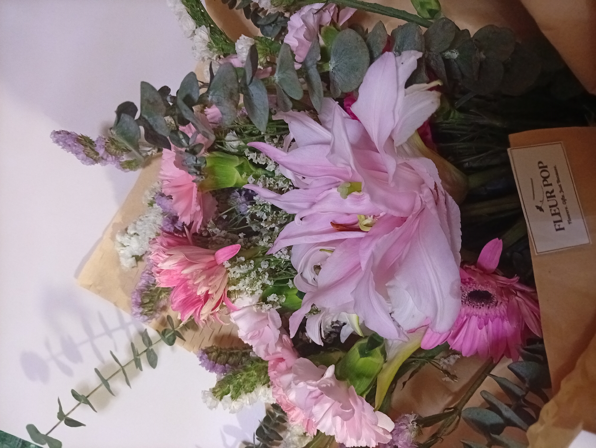 The image displays a bouquet of pink and white flowers, likely a gift. The arrangement features lilies, carnations, and other smaller filler flowers, wrapped in brown paper with a "FLEUR POP Flowers, Gifts, Just Because" label. The flowers are resting on a light-colored surface. The image does not contain enough information to determine the specific location in Davao City, Philippines.