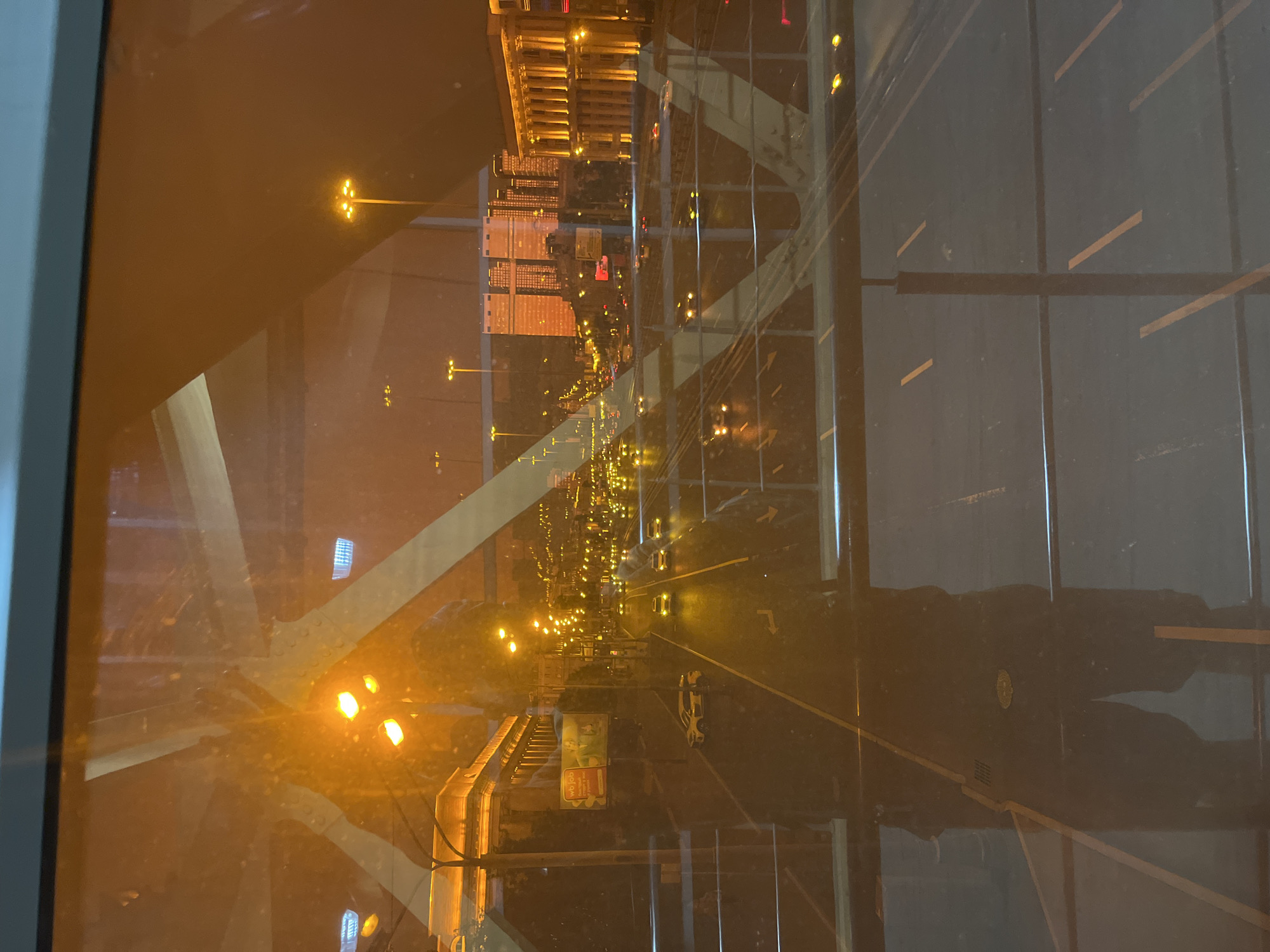 The image shows a reflection in a window, revealing a nighttime cityscape. In the foreground, a person's reflection is visible, standing on what appears to be an elevated walkway or bridge. The person is wearing dark clothing. Beyond the reflection, a multi-lane road is visible with several cars driving on it, their headlights and taillights illuminated. Streetlights line the road, casting a warm glow. In the background, tall buildings with illuminated windows are present. A billboard with an advertisement is also visible on the side of a building. The overall scene suggests an urban environment at night. This is a reflection of the Leningradskoye Highway in Khimki, Russia.