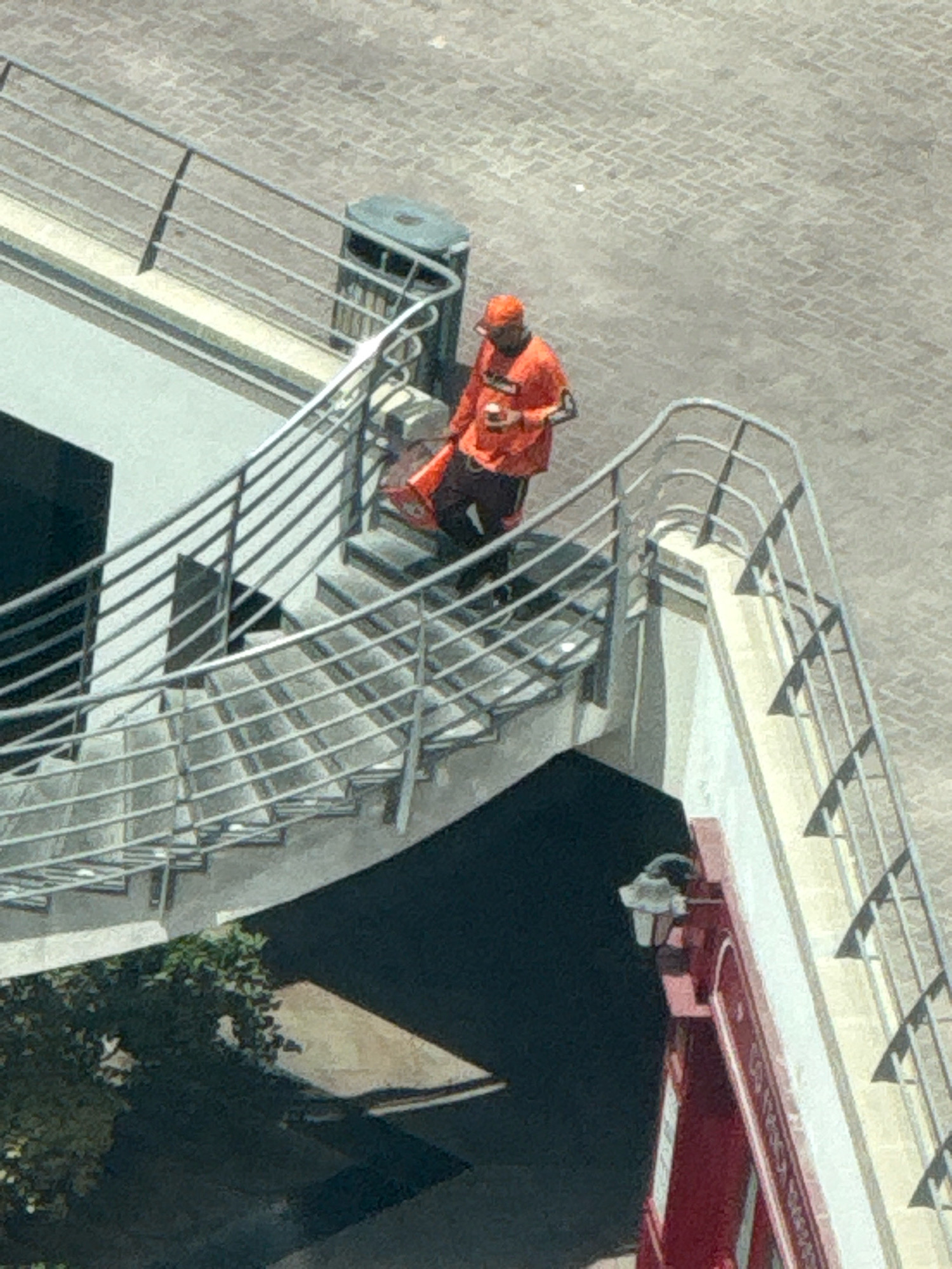 A person wearing an orange jacket and cap is standing on a concrete staircase with metal railings. The person is holding a red bag. The staircase appears to be outdoors, with a paved surface in the background. There is a trash bin next to the top of the staircase. A building with dark windows is visible on the left side of the staircase. A red structure with a sign is partially visible on the right side of the staircase. The overall scene suggests an urban environment, possibly in Dubai, United Arab Emirates, given the architectural style and paving.