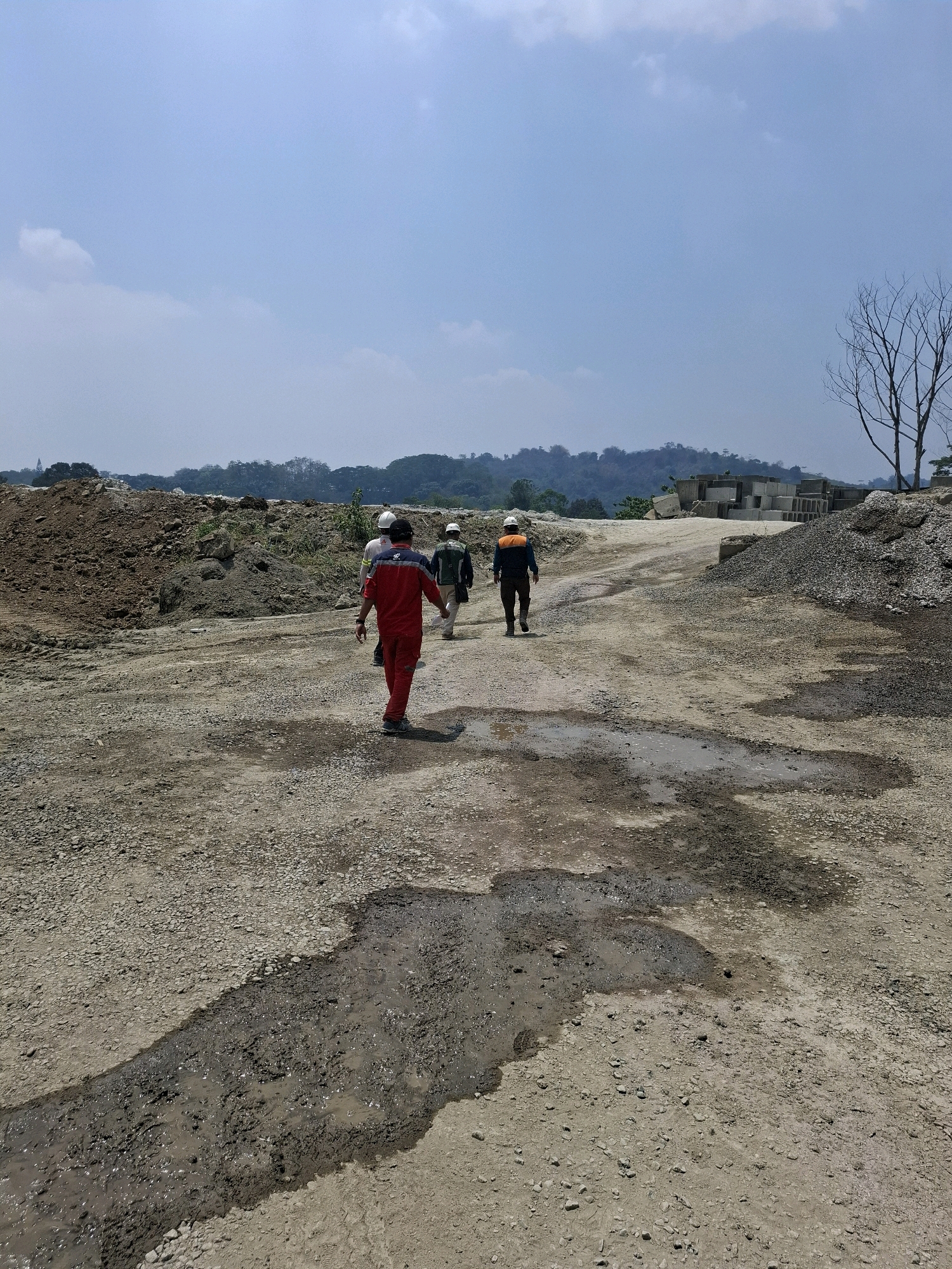 Four individuals, three appearing male, walk along a dirt and gravel road within a construction or quarry site. The person in the foreground wears a red and blue uniform with a white hard hat. To their left, another person in a white hard hat and light-colored shirt walks. Further ahead, two more individuals are visible; one wears an orange vest and dark pants, and the other, further to the right, wears a backpack and dark pants with a light-colored shirt. Puddles of muddy water are present on the road. The environment is arid, with piles of dirt and rock, concrete blocks, and sparse vegetation in the background. A bare tree stands to the right. The sky is mostly clear with scattered clouds. The scene suggests an industrial or developmental area, potentially in or near Cijangkar, Indonesia, based on the provided context.