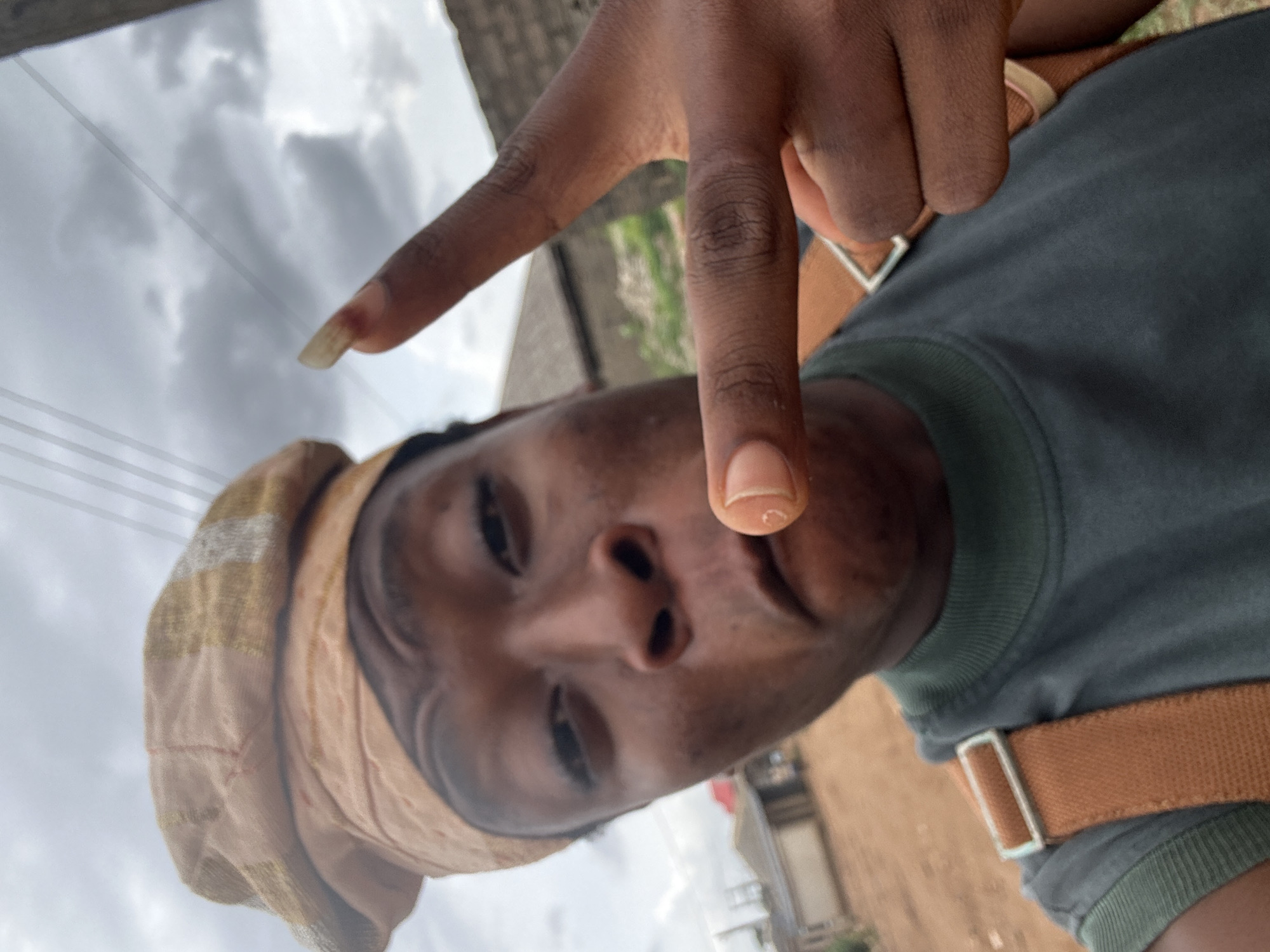 A person poses for a selfie outdoors. They are wearing a tan headscarf and a gray-green shirt with brown straps, possibly from a backpack or bag. The person's hand is raised, making a "V" sign towards the camera. In the background, there are buildings and utility lines against a cloudy sky. The location appears to be urban.