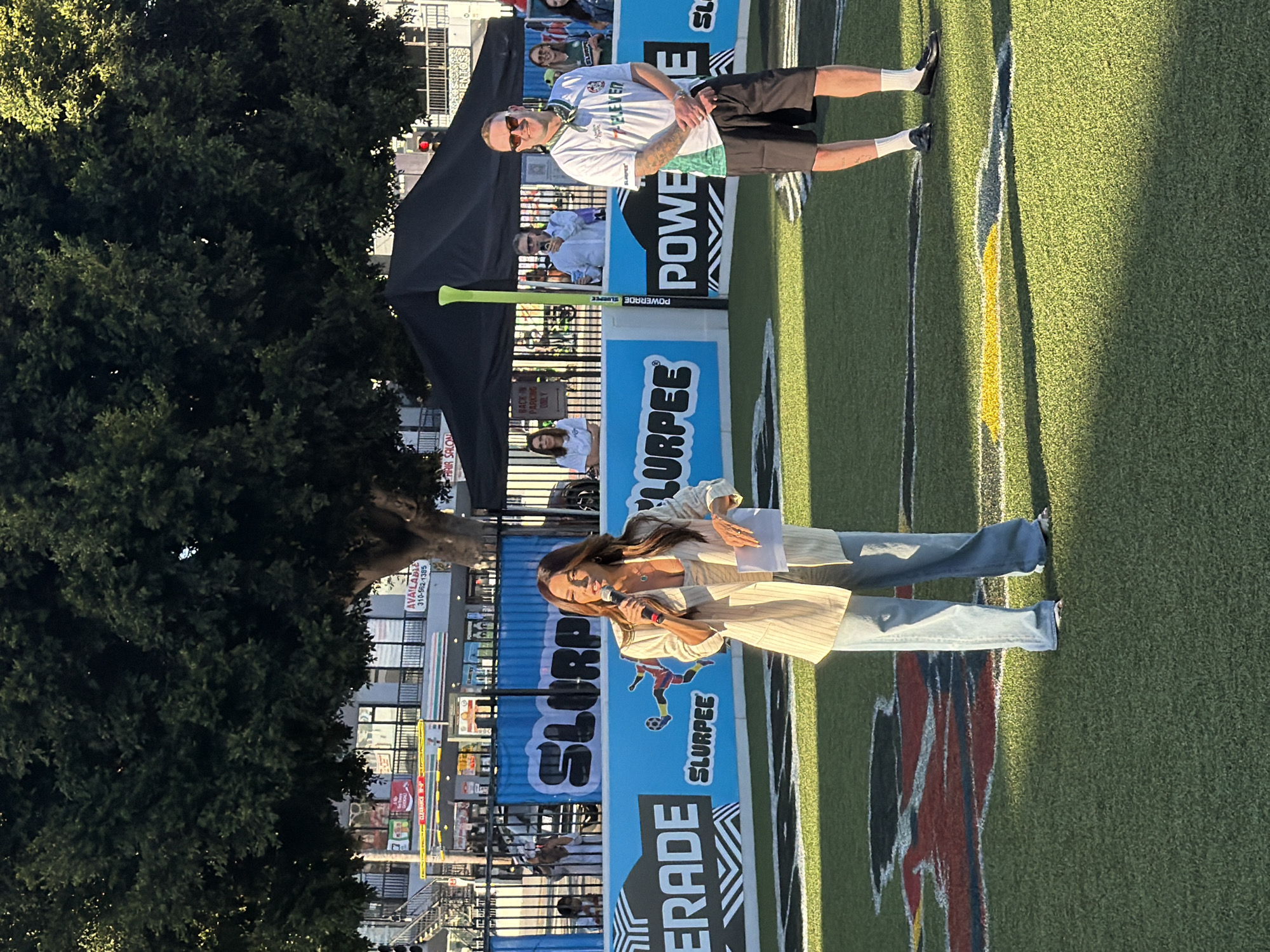 The image depicts an outdoor event on a synthetic turf field, likely a sports field. The backdrop features a blue banner with "SLURPEE" branding and graphics of soccer players. To the left, "GATORADE" is visible on a similar banner. The event appears to be sponsored by Slurpee and Gatorade.
In the foreground, a woman is speaking into a microphone, holding a piece of paper. She is wearing a light-colored pinstriped blazer over a white top and light-wash denim jeans. To her right stands a man wearing sunglasses, a white t-shirt with green and black accents, black shorts, white socks, and black loafers. He has tattoos on his arms.
In the background, there are people standing behind a fence. A large tree with dense foliage dominates the upper left portion of the image, casting shadows on the scene. Behind the tree, buildings are visible, including a "7-ELEVEN" store with its characteristic green and orange signage. The overall setting suggests a public event or promotion in an urban environment. The presence of a 7-Eleven store and the style of the buildings hint at a location within the city of Los Angeles, United States of America.