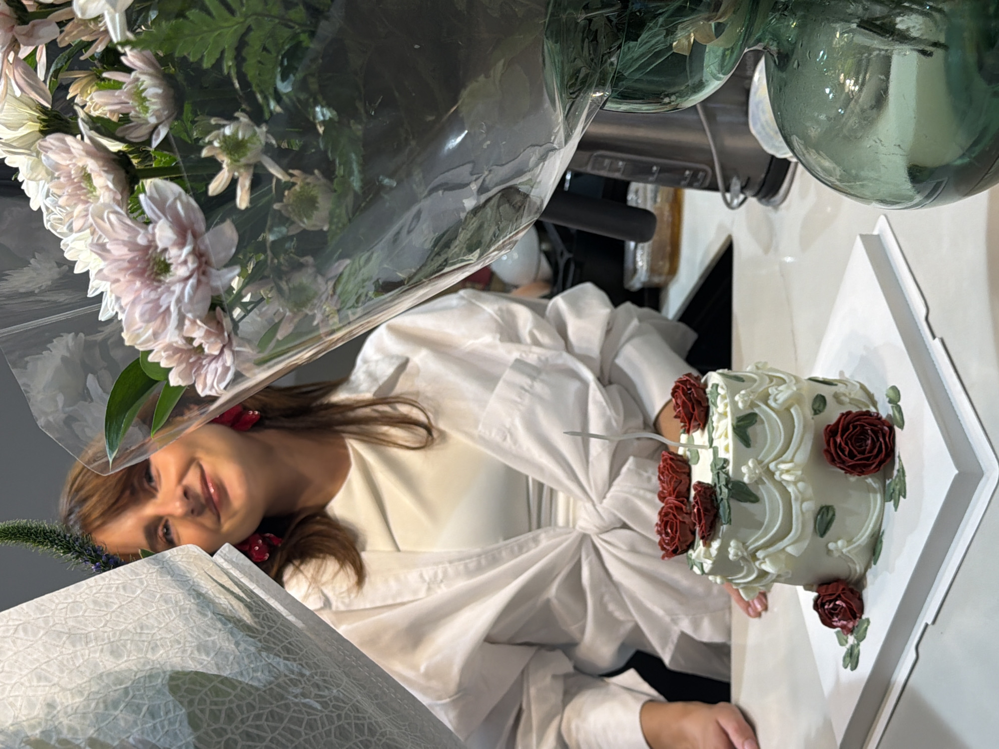 A woman is seated at a table, with a small cake and a bouquet of flowers in front of her. The woman is wearing a white shirt and has long brown hair. The cake is white with red roses. The flowers are pink and white. The scene appears to be indoors, possibly a home or restaurant. The image is taken at a slight angle, with the woman's face and upper body visible.