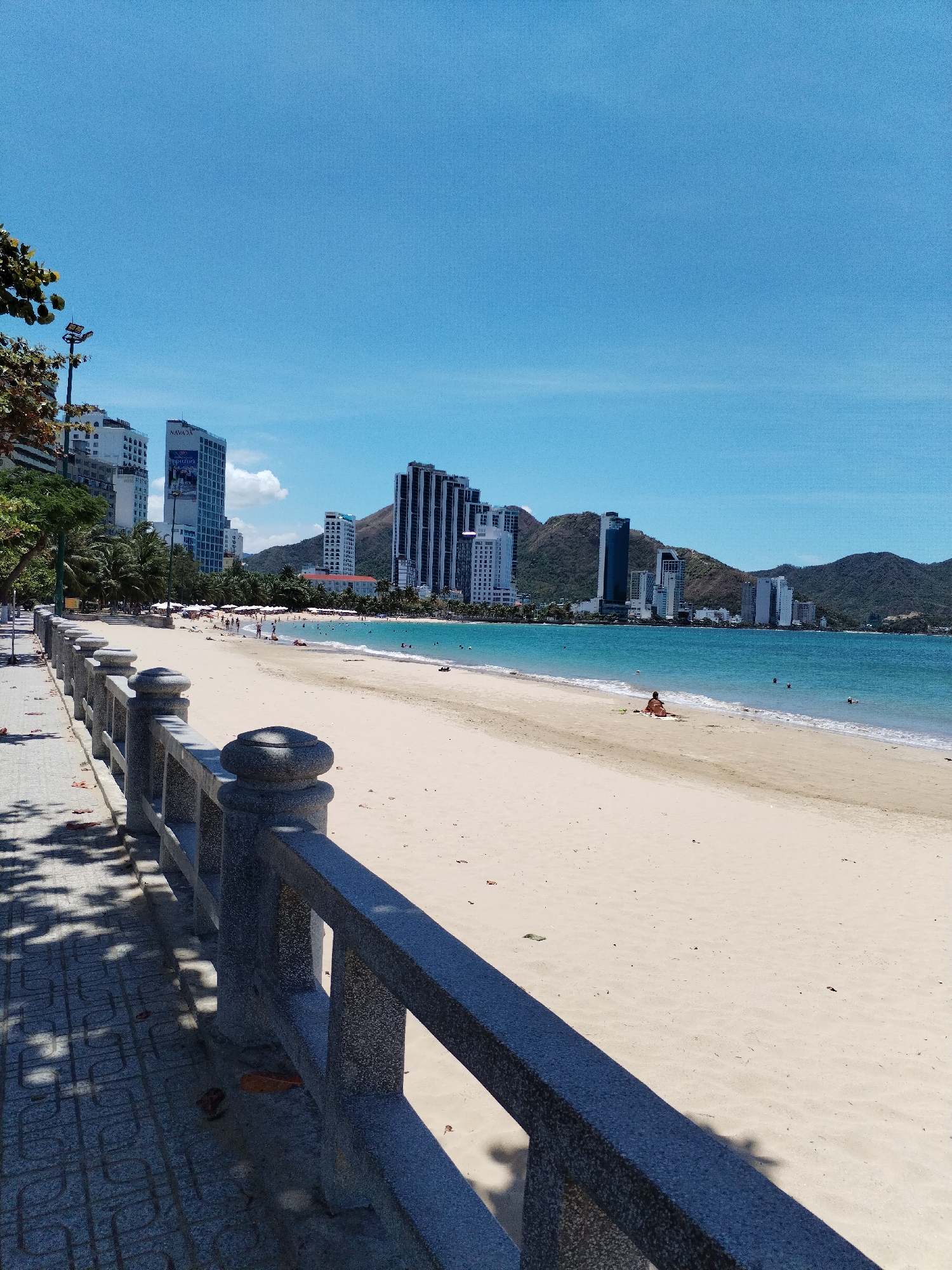 The scene depicts a beach in Nha Trang, Vietnam, characterized by a light sandy beach and turquoise water. In the background, modern high-rise buildings blend with verdant hills. A small number of people are scattered across the beach, some appearing to be sunbathing and others swimming. A granite-style railing is on the left side of the image. The sky is clear and blue.
