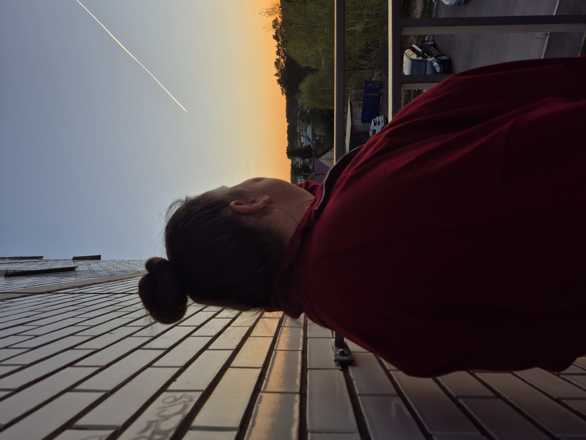 A low-angle, vertically-oriented shot captures a woman looking toward a sunset sky, likely from a balcony in Chaikovsky, Russia. The sky displays a gradient, transitioning from blue to orange near the horizon, with a contrail stretching across. The woman, wearing a red garment, is partially visible, with her head tilted upwards and her hair styled in a bun. A tiled wall extends from the lower left corner towards the center. In the background, beyond the balcony railing, are trees and rooftops, characteristic of a residential area. Several vehicles are visible in the background on the right. The image is shot at dusk or dawn, as indicated by the lighting.