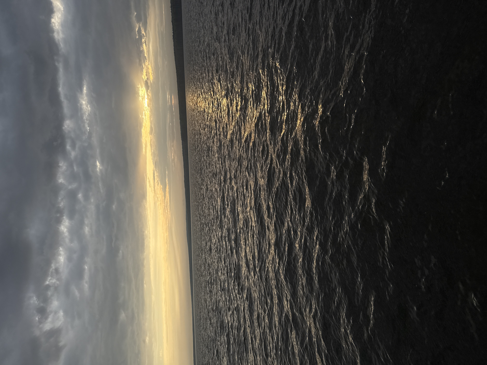 The image shows a body of water, likely the Kama River, at sunset or sunrise. The sky is filled with dark, textured clouds on the left, transitioning to a lighter, golden hue where the sun is breaking through. The water surface reflects the sunlight, creating a shimmering effect. A thin strip of land is visible on the horizon. The scene has a calm, serene atmosphere.
