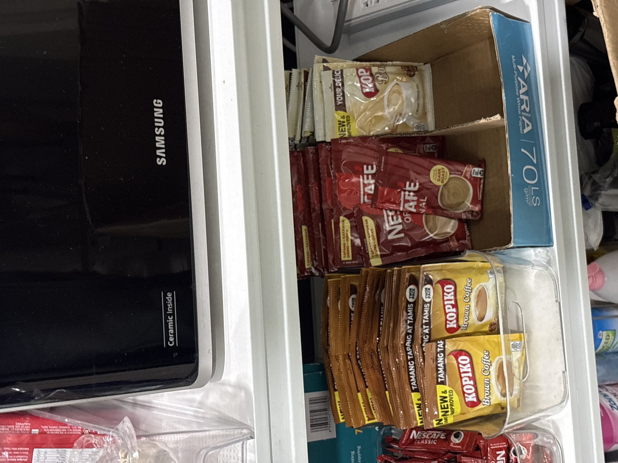 The image shows a shelf filled with various coffee products. In the foreground, on the left, a clear plastic container holds several sachets of Kopiko Brown Coffee. To the right of the container, a cardboard box is filled with more coffee sachets, including Nescafe and Kopiko brands. In the background, a black Samsung microwave with "Ceramic Inside" printed on it is visible. The overall environment appears to be a kitchen or storage area. The presence of specific coffee brands and the microwave suggests a domestic setting. There are no people visible in the image.