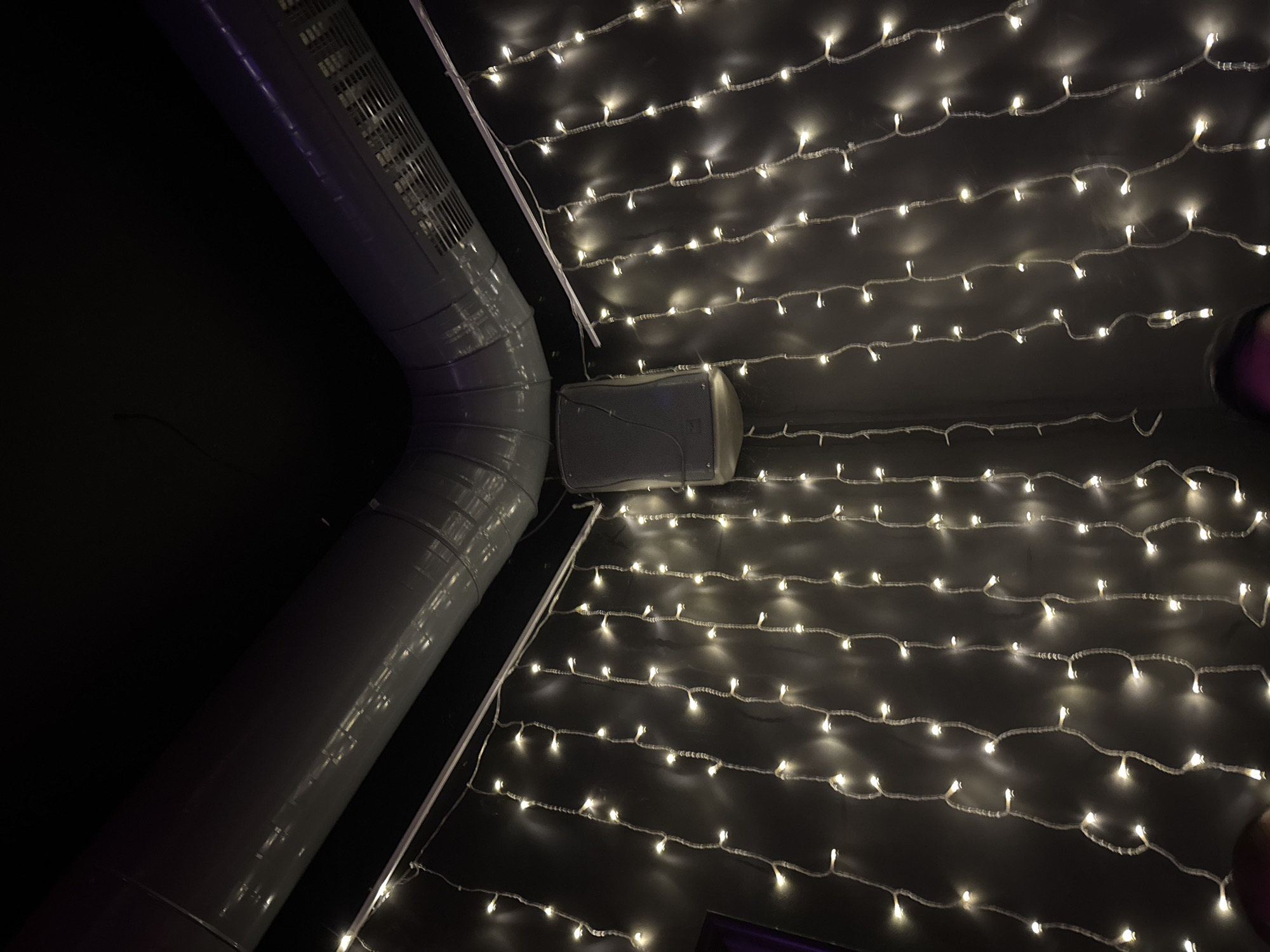 The image depicts an upward-angled view into the corner of a dimly lit interior space. A large, grey, industrial-style ventilation duct system runs horizontally across the upper portion of the frame, bending at a right angle in the center. The right segment of the duct features a visible vent grille. Below the ductwork and mounted on the left wall, a grey, wedge-shaped speaker with a textured grille is visible. The dark walls are adorned with numerous vertical strands of warm white LED string lights, suspended from a horizontal white track or rail located just below the ductwork. These lights provide the primary illumination for the scene. At the very bottom edge of the frame, the top of a dark-haired human head, showing a subtle purple reflection on the left side, is partially visible. The specific location within Sofia, Bulgaria, cannot be determined from the visual evidence.
