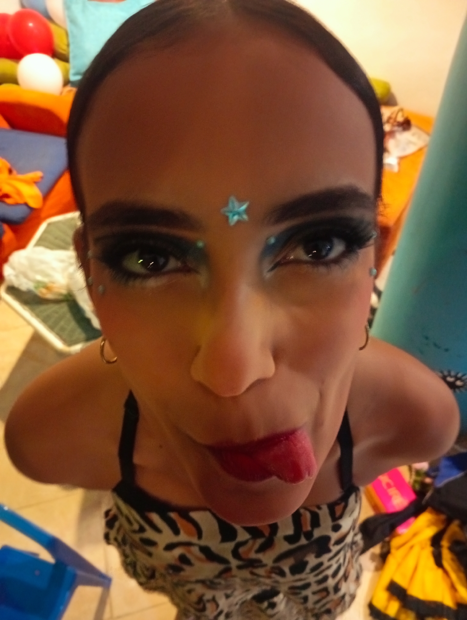 A close-up, high-angle shot shows a person with brown skin and dark hair pulled back, sticking out their tongue. They are wearing heavy eye makeup with blue accents, a blue star on their forehead, and small blue dots near their eyes. Gold hoop earrings are visible. The person is wearing a patterned top with thin black straps.
The background includes a variety of objects such as pillows, balloons, a small table with items on it, a blue cylindrical object, and various clothing items. The setting appears to be indoors, cluttered with personal belongings. There is no verifiable evidence to place this image specifically in Playa El Agua, Venezuela.
