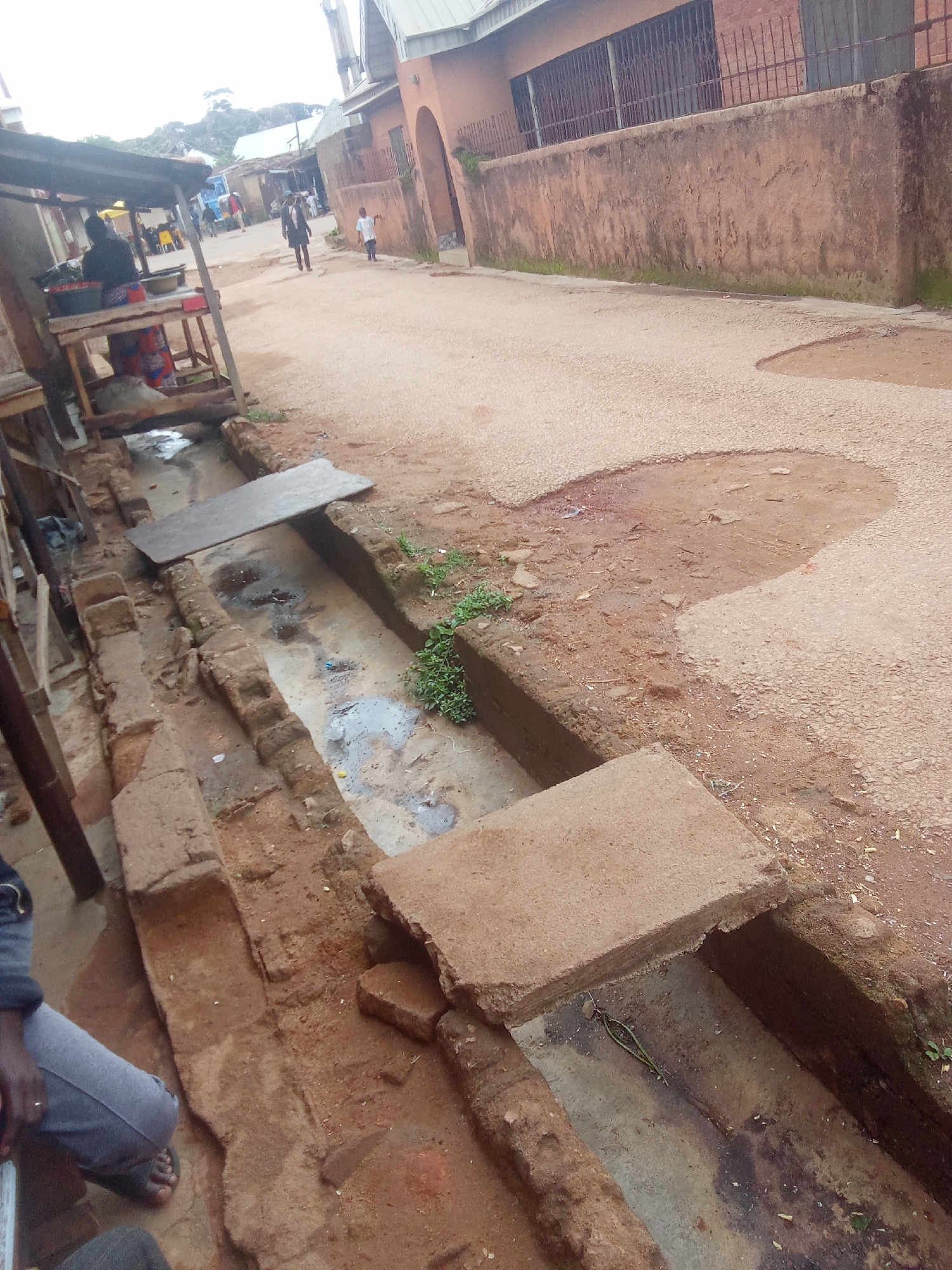 A ground-level, medium-distance, eye-level shot shows a street scene in Jos, Nigeria. In the foreground, an open, brick-lined drainage channel runs horizontally across the image. The channel is partially covered by flat concrete slabs, with one slab in the center of the frame and another to the right. The channel appears to contain murky water and debris. A person wearing grey trousers and sandals is partially visible in the lower left corner of the image, kneeling beside the channel.
To the left of the channel, a makeshift wooden stall with a corrugated metal roof is set up. A person wearing a dark top and a patterned skirt stands behind the stall, appearing to be attending to goods displayed on the shelves.
The street itself is unpaved and appears dusty, with some potholes and patches of uneven terrain. In the background, buildings with textured walls line the street, and some individuals are visible walking along the road. The overall environment suggests a bustling, informal urban setting.
