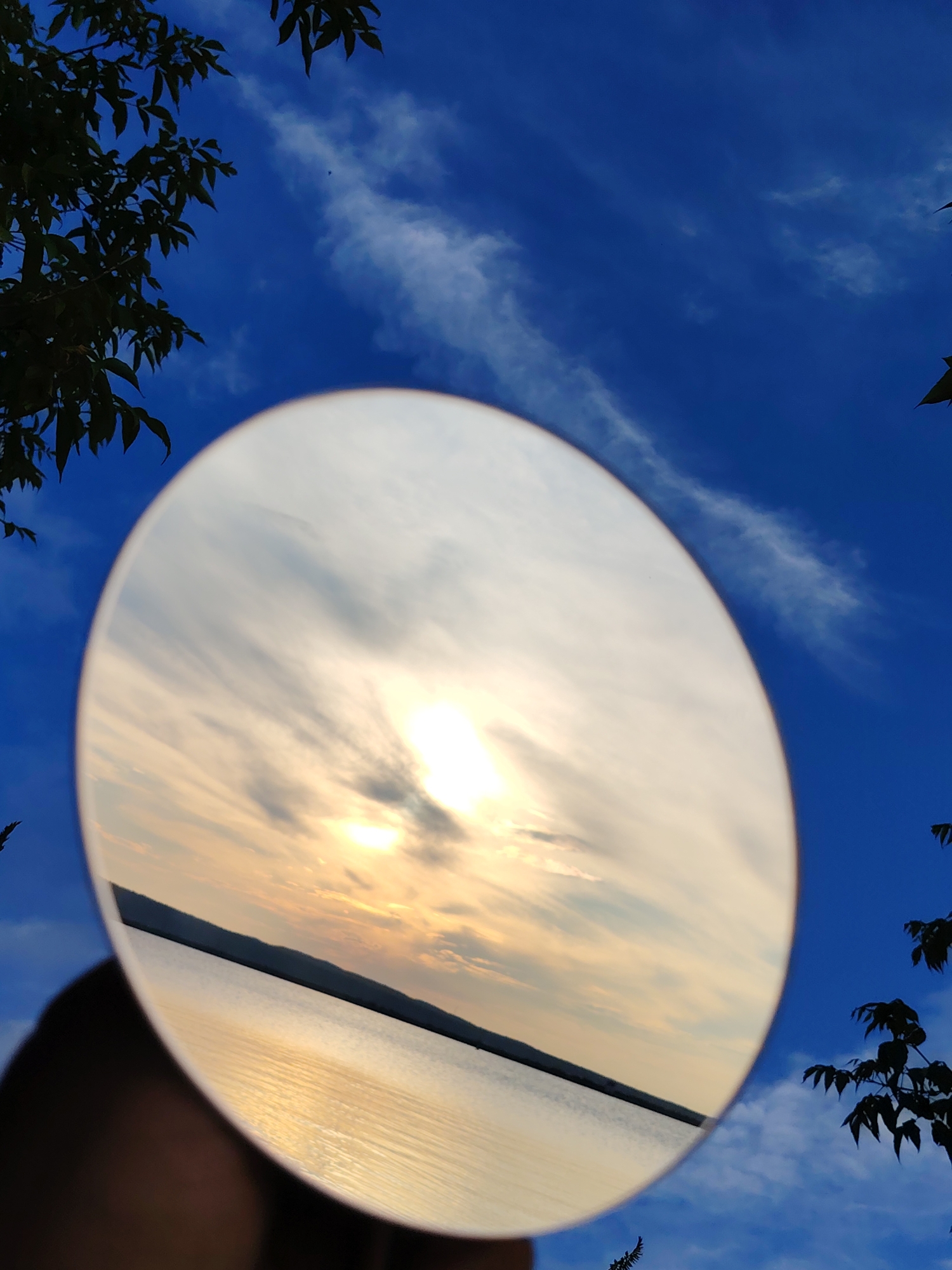 The image is a close-up, upward-angle shot of a mirror reflecting a sunset over a body of water, set against a bright blue sky with wispy clouds. Silhouetted tree branches are visible in the foreground and along the edges of the frame, extending from the top left and right. The mirror itself is circular and held up by an unseen hand, partially obscuring the sky. The reflection shows a calm body of water with a dark, silhouetted horizon line, likely a distant shore or hills. The sun is a bright, blown-out orb in the reflection, surrounded by streaks of orange, yellow, and white clouds. The overall impression is one of nature and reflection, with the mirror acting as a window to a different view. The location is identified as Chaikovsky, Russia, implying the scene takes place near a body of water relevant to the city, such as the Kama River or a reservoir.