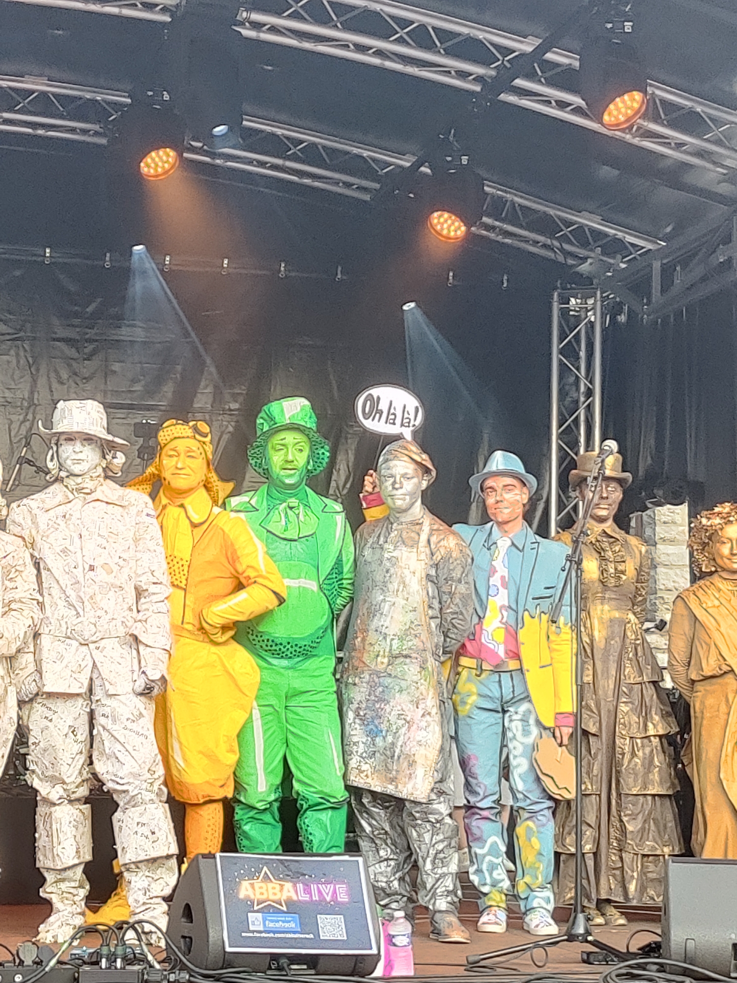 The image depicts a performance stage in Marche-en-Famenne, Belgium, where a group of individuals dressed as statues are gathered. The performers are adorned in full body paint, with one wearing a white costume resembling old paper, another in a bright yellow outfit with a pilot's helmet, and a third in a vibrant green suit and hat. To the right, additional performers are dressed in metallic gold and bronze costumes, one with an elaborate layered dress and another in a toga-like garment. A speech bubble saying "Oh la la!" hovers above one of the performers. In the foreground, a speaker with an "ABBALIVE" logo is visible, along with parts of audio equipment. The stage is illuminated by spotlights, creating a dramatic effect against a dark backdrop. The overall scene suggests a street performance or an outdoor event.