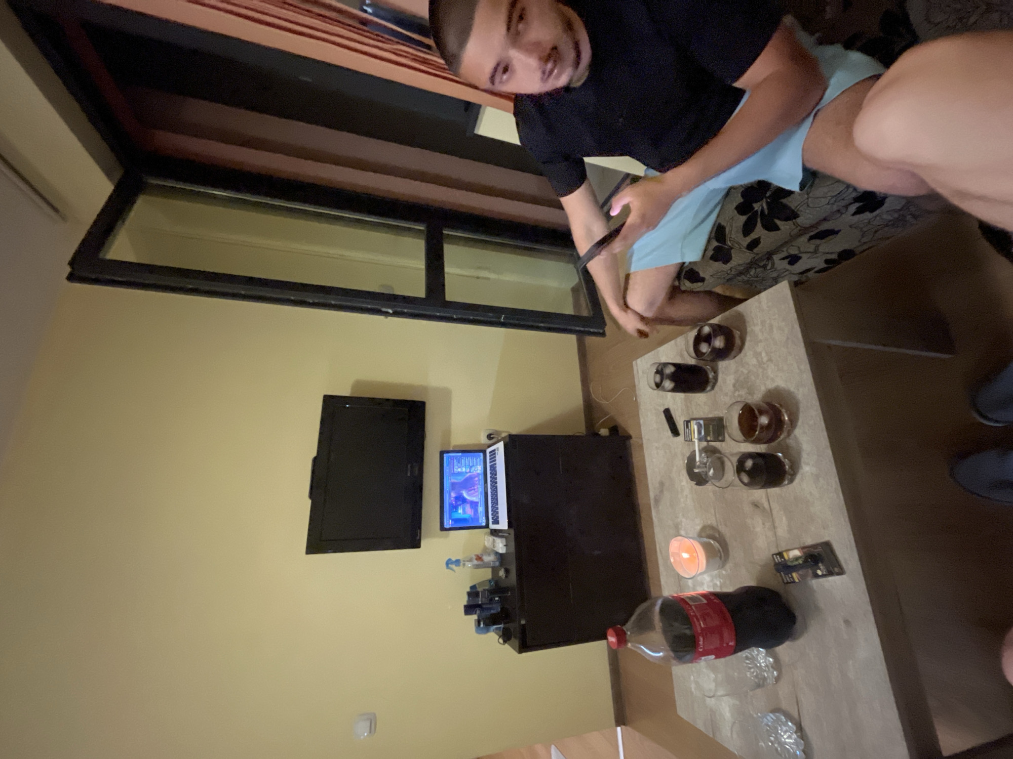 The image depicts a room in Plovdiv, Bulgaria. A person wearing a black shirt and light blue shorts sits on a patterned couch, holding a phone. A coffee table in front of the couch holds glasses, a bottle of Coca-Cola, a candle, and other small objects. In the background, there's a TV mounted on the wall, along with a computer setup that includes a monitor and a computer case. A large window is also visible.