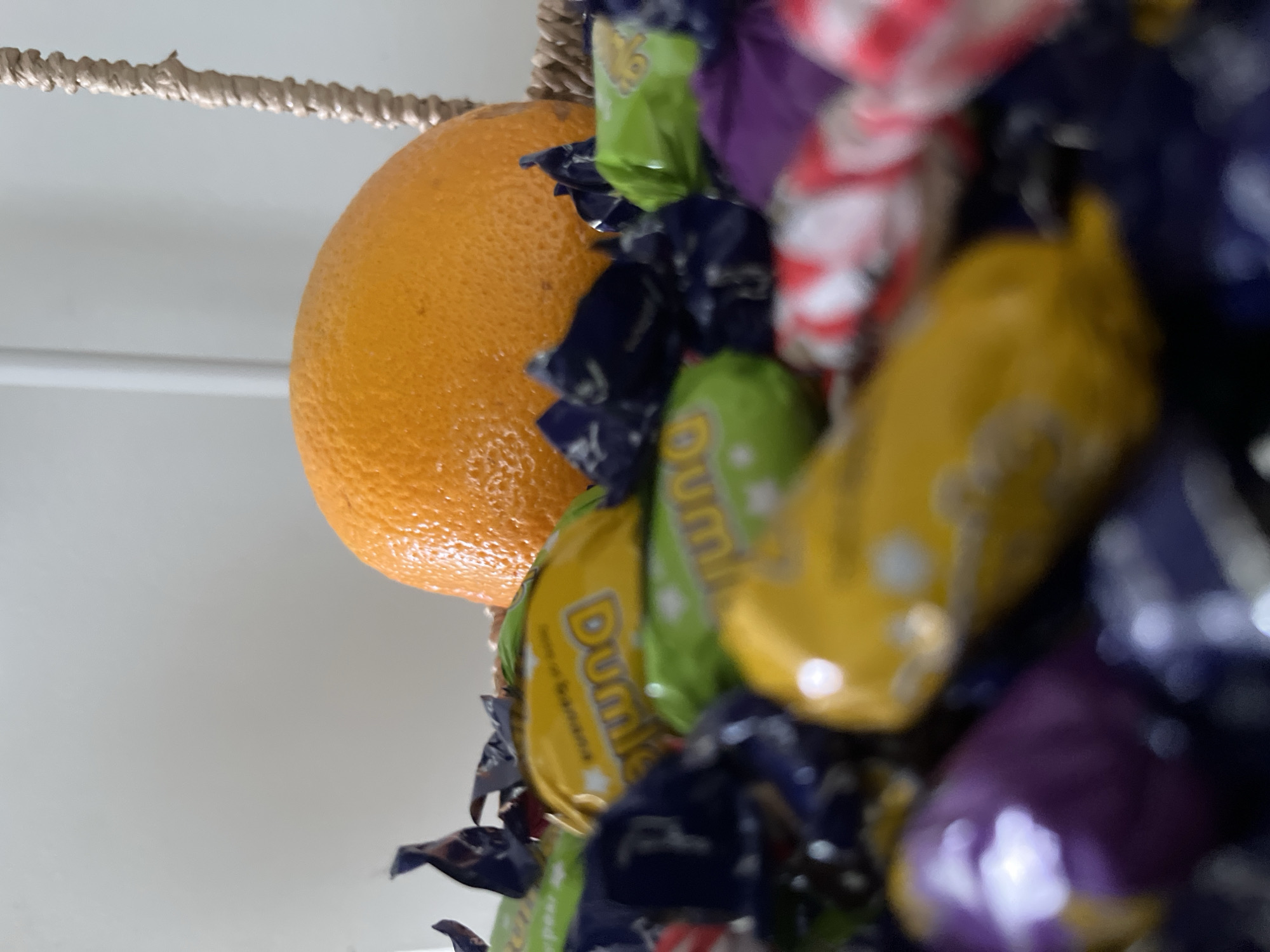 An orange sits in a woven basket filled with individually wrapped candies. The orange is in the upper right quadrant of the image. The candies are in various colors, including yellow, green, purple, and red and white striped wrappers. Some of the yellow and green wrappers appear to have the word "Dumle" printed on them. The basket is partially visible on the right side of the image, and a light-colored wall is in the background. The scene is brightly lit, and the focus is on the candies and the orange. There are no people or animals present. The location is not specified beyond the context of being in a home or shop in Helsinki, Finland.