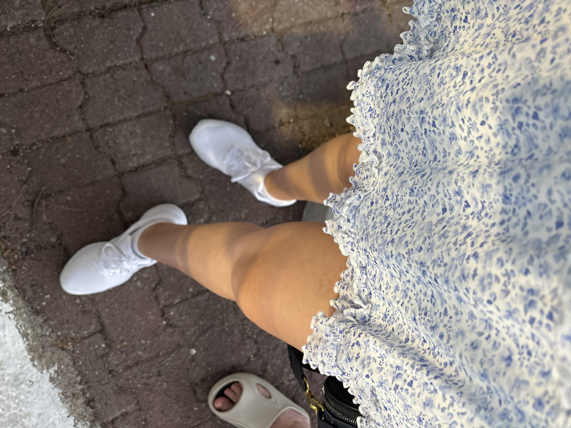A high-angle shot depicts a person standing on a paved surface, likely a pedestrian walkway. The person is wearing a short, light-colored dress with a blue floral pattern and white sneakers. Another foot, wearing a slide sandal, is partially visible in the bottom left corner, suggesting the presence of another individual. The surface appears to be made of interlocking pavers. The location appears to be outdoors. Without specific landmarks or contextual details, a precise location within Budva, Montenegro, cannot be determined from this image alone.