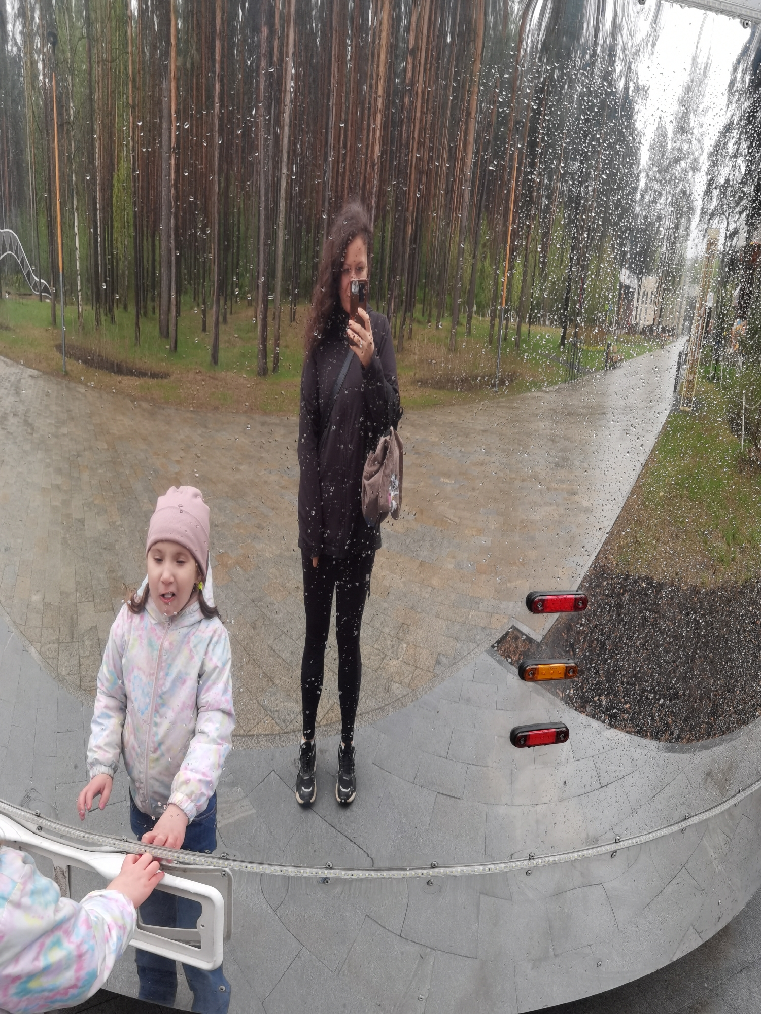 The image, taken in Perm, Russia, shows the reflection of a woman and two children in a large, curved, metallic surface, likely a public art installation or feature. The woman is standing and holding a mobile phone, possibly taking the picture. She has dark curly hair and is wearing a black hoodie, black leggings, and black sneakers. A small bag hangs from her shoulder. One child is a young girl in a pink hat and patterned jacket, touching the surface. Another child's arm and hand are visible touching the surface as well. In the reflection, behind the subjects, there is a forest of tall, slender trees and a walkway. The surface is wet with raindrops, and the image includes the reflection of two small, horizontally-oriented lights: one red, one amber. The ground in the reflection appears to be paved.