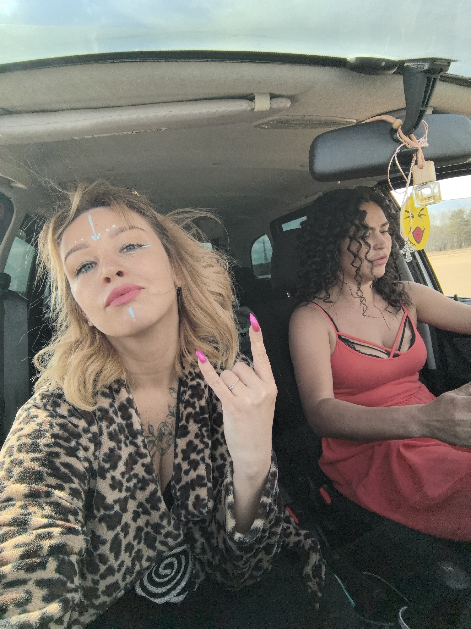 Two women are captured in a selfie taken inside a car during what appears to be daytime with clear, sunny weather, potentially in Chaikovsky, Russia. The woman on the left, who is taking the photo, has blonde hair and light-colored eyes. She wears a leopard print robe or jacket and has distinctive light blue makeup, including an arrow and dots on her forehead and a line on her cheek. Her lips are painted with pink lipstick, and she makes a "rock on" hand gesture with her nails painted bright pink. A tattoo featuring floral elements is visible on her chest/shoulder area.
The second woman, seated in the driver's seat to the right, has dark, curly hair and wears a red top or dress with black criss-cross straps. She looks away from the camera, towards the right side of the vehicle, her expression appearing neutral or thoughtful.
The car's interior is visible, including a light-colored headliner and a rearview mirror. Hanging from the rearview mirror is a yellow, smiley-face-shaped air freshener and a smaller, clear bottle air freshener. The word "Freon" is visible on the yellow air freshener. Through the windshield, a clear blue sky is visible, and in the distance, there appears to be an open field or roadside with trees. Both women seem relaxed within the car.