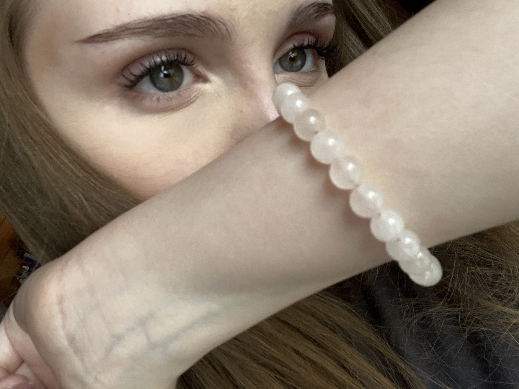 The image displays a close-up of a person's face and arm, with a pearl bracelet on the arm. The person has light skin, brown hair, and green eyes, and appears to be wearing makeup. The background is dark and out of focus. There is no information in the image that would suggest a location in the city of Ashkadar, Russia.
