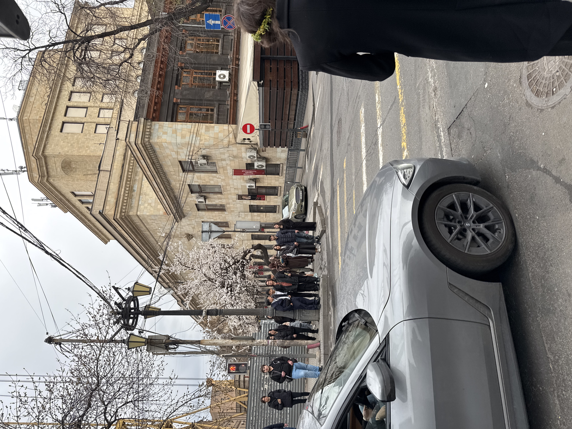 A medium, eye-level shot depicts a street scene in Yerevan, Armenia. A gray car dominates the foreground, with its front tire occupying the right side of the frame. A group of people are standing along a sidewalk to the left, gathered near a flowering tree with white blossoms. Behind them, a multi-story stone building is visible, along with air conditioning units affixed to its facade. Traffic signals and street lamps are present. Overhead electrical wires crisscross the sky. The sky is overcast. A person wearing a floral crown is standing near a street sign.