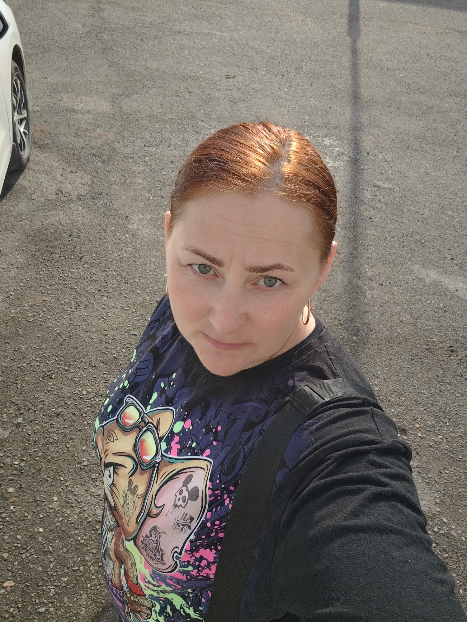 A woman with red hair and blue eyes is taking a selfie outdoors on an asphalt surface, likely a street or parking lot. A portion of a white car is visible to the upper left. The woman is wearing a black t-shirt with a colorful graphic design, featuring a cartoon-like figure. She has a black strap, possibly from a bag, across her shoulder. She wears gold hoop earrings. The location is in Markovskii, Russia.