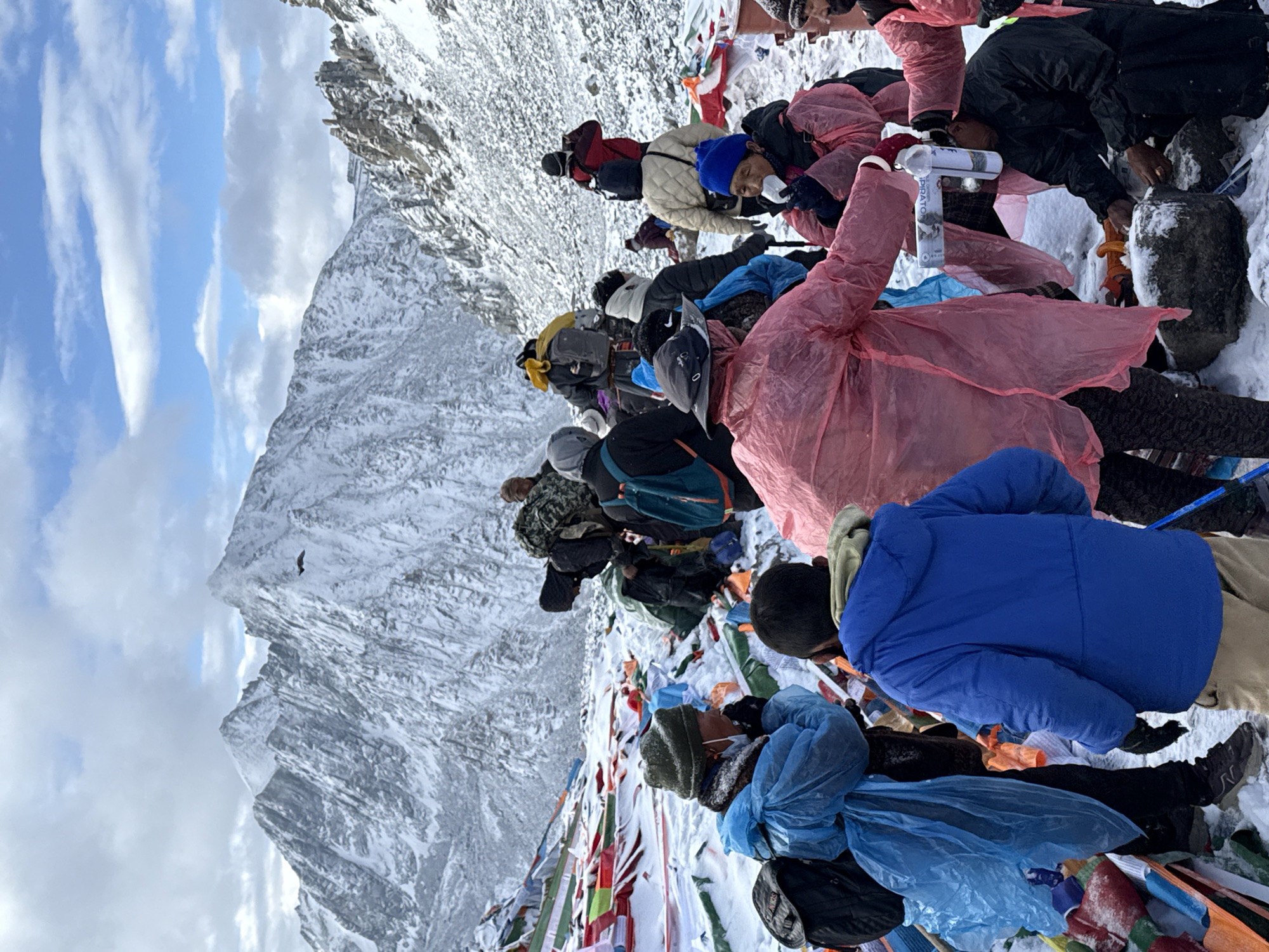 This image captures a group of people gathered on a snowy mountain slope, likely during a pilgrimage or trek in a high-altitude region near Parga town, China. The scene is set against a backdrop of majestic, snow-covered peaks under a partly cloudy sky with visible blue patches.
Numerous individuals are visible, dressed in heavy winter clothing. Many are wearing colored rain ponchos—prominently pink and blue—over their jackets, suggesting protection from wind, snow, or rain. They appear to be resting on the snowy ground, with some huddled close together. Colorful prayer flags (Lungta) are extensively draped across the snow-covered rocks and ground, indicating a sacred or culturally significant location.
Notable objects include backpacks worn by several people. One person in a pink poncho is holding a white plastic bottle, partially labeled "HYDRATOR" and "DRINK," suggesting rehydration during their journey. Another person appears to be handling a walking pole or ice axe. In the upper left portion of the image, a small bird is visible in flight against the snowy mountainside.
The activity taking place is a rest stop amidst a challenging high-mountain environment. The presence of prayer flags strongly implies a spiritual or cultural pilgrimage. The time of day is clearly daytime, and the weather is cold and snowy. The overall impression is one of endurance and devotion in a stunning, yet harsh, natural setting.
