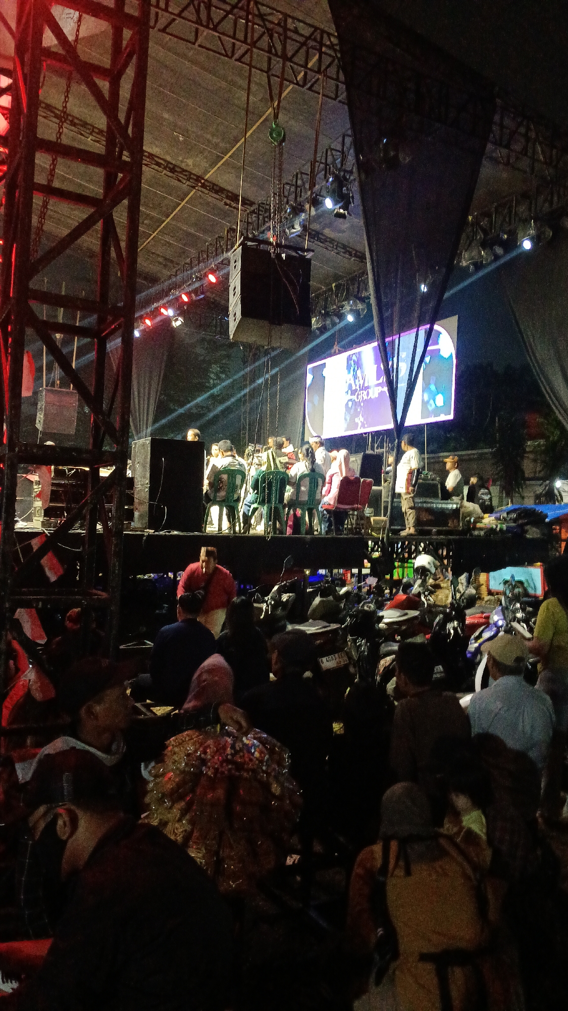 The image depicts an outdoor event at night in Tajurhalang, Indonesia, likely a concert or festival. A stage is set up with a large screen displaying a logo that reads "AMLY GROUP". There are multiple speakers suspended from a metal truss structure above the stage. Several individuals are seated on the stage, some appearing to be performers or organizers. In the foreground, a crowd of people is gathered, many of whom are seated or standing around parked motorcycles. The lighting is dim, with spotlights illuminating the stage and the screen. The overall atmosphere suggests a lively event taking place in a public space.