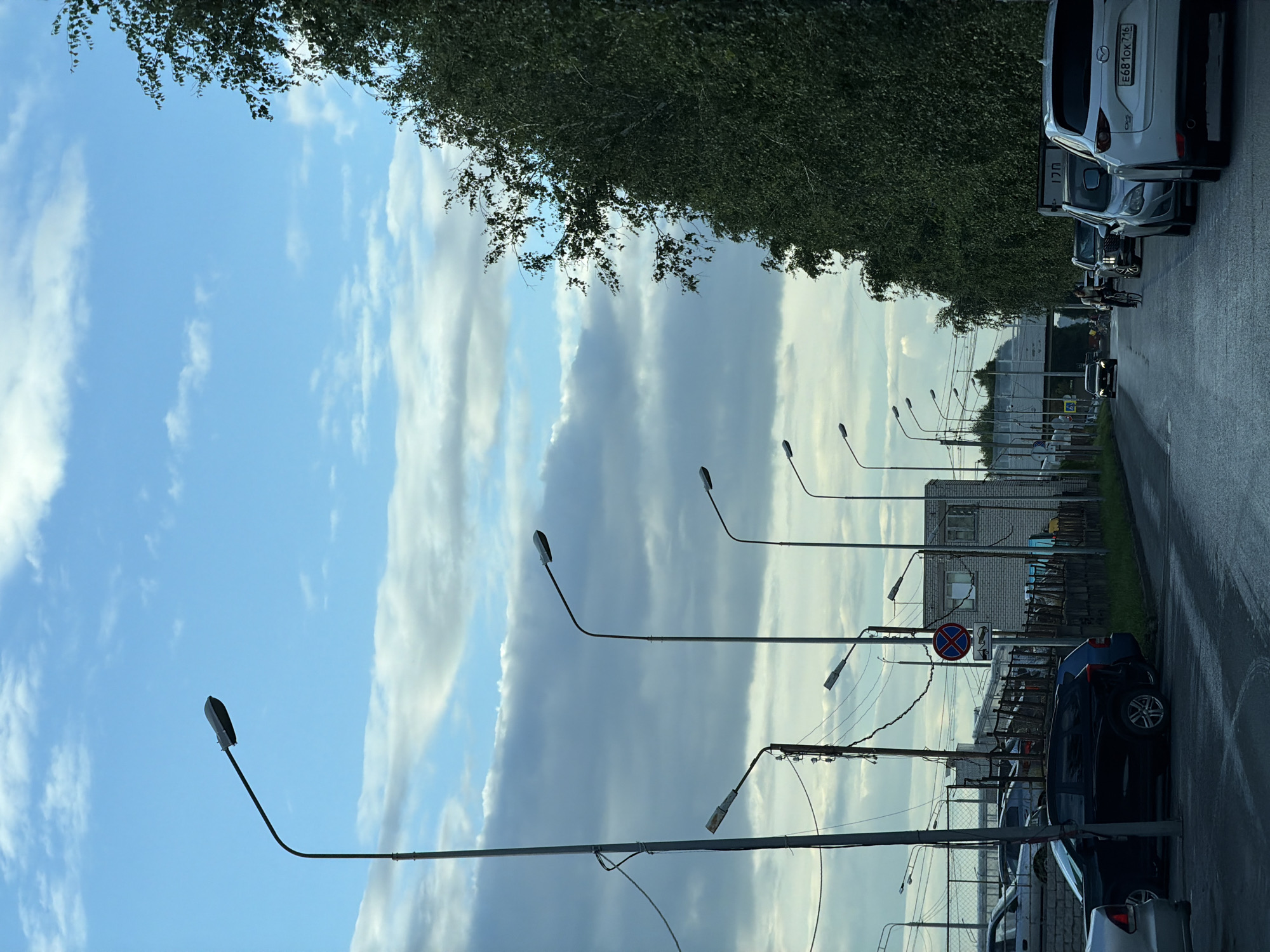 The image depicts a street scene in Kazan, Russia, under a cloudy sky. Several tall streetlights line the left side of the road. In the foreground, a black SUV is parked, and other cars are visible further down the street. On the right, a dense line of trees forms a natural barrier. A white SUV with the license plate E681ok 716 is parked on the right side of the road. There are buildings and a fence in the background. A person is seen riding a bicycle further down the street, and other vehicles are present in the distance. The road appears to be wet, suggesting recent rain. A traffic sign indicating "no stopping" is visible on the left side of the road.
