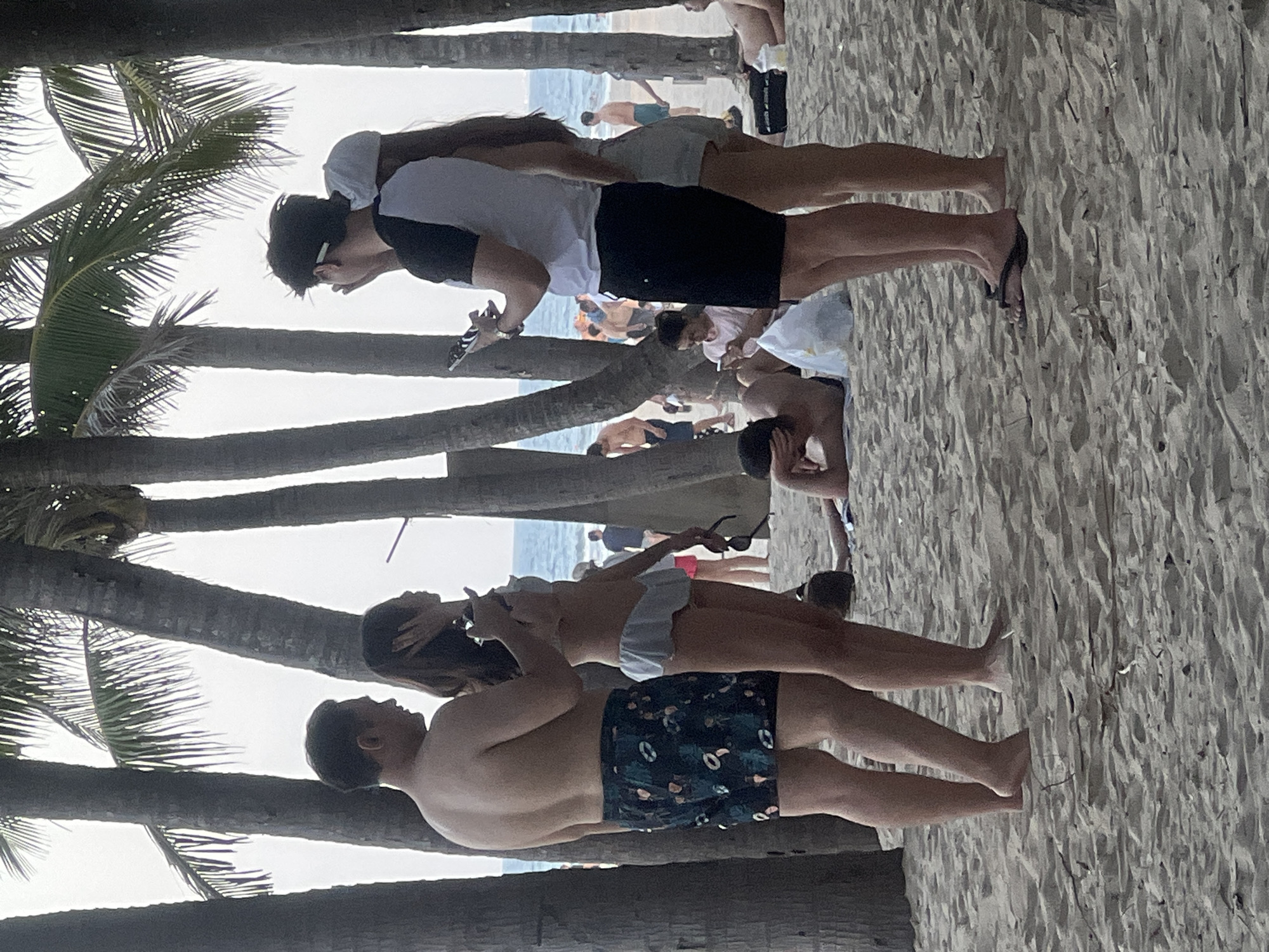 At a beach in Da Nang, Vietnam, a group of people are gathered. In the foreground, a man in patterned swim trunks stands next to a woman in a light blue bikini. The man is shirtless, and the woman has her hand near her face. To their right, another couple stands, with the man wearing a black and white shirt and black shorts, and the woman wearing a light-colored hat and shorts. The man is holding a phone. Further back, other beachgoers are visible, some sitting or lying on the sand, and others standing near the water. Palm trees line the beach, and the ocean is visible in the background. The sand is a light brown color.