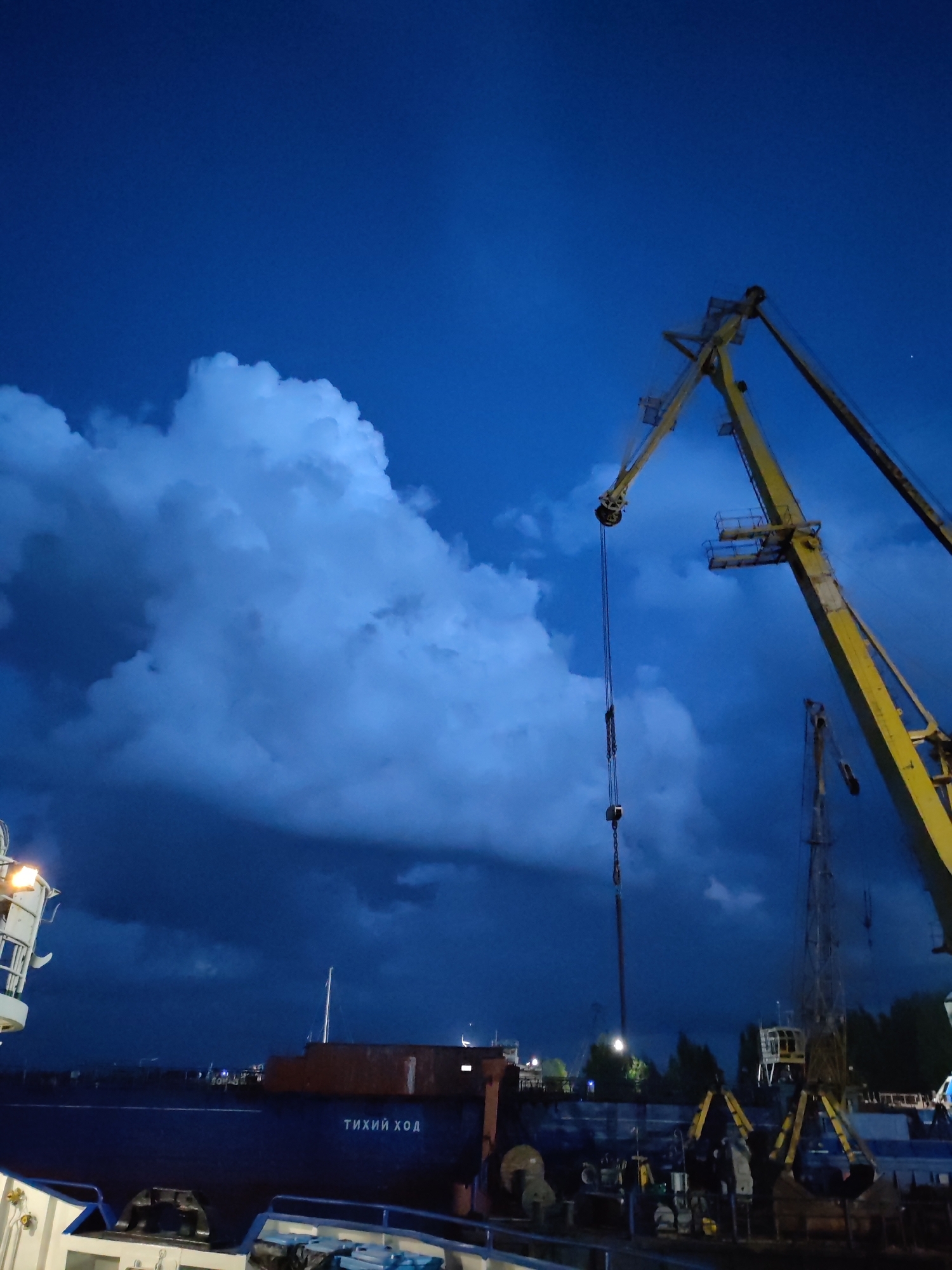 The image shows a dark blue sky with dramatic, illuminated clouds over a port area in Rybinsk, Russia. A large yellow crane dominates the right side of the frame, its boom extending upwards. Below, a dark blue ship with the Cyrillic text "ТИХИЙ ХОД" (Tikhiy Khod, meaning "Slow Speed") on its hull is docked. Other port machinery, including smaller cranes and equipment, are visible in the background. The overall lighting suggests it is either twilight or nighttime, with some artificial lights illuminating parts of the scene. There are no people clearly visible in the image.