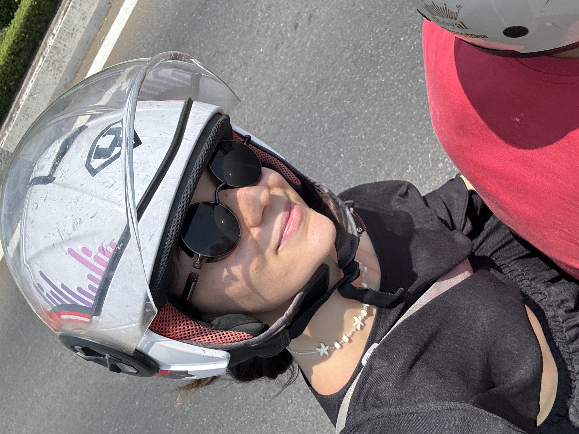 A woman with sunglasses is wearing a motorcycle helmet. She is riding on a road next to a person wearing a red shirt and a white helmet that says “Royal”. The location is likely Phú Thủy, Vietnam. The woman wears a necklace made of white flowers and stars.
